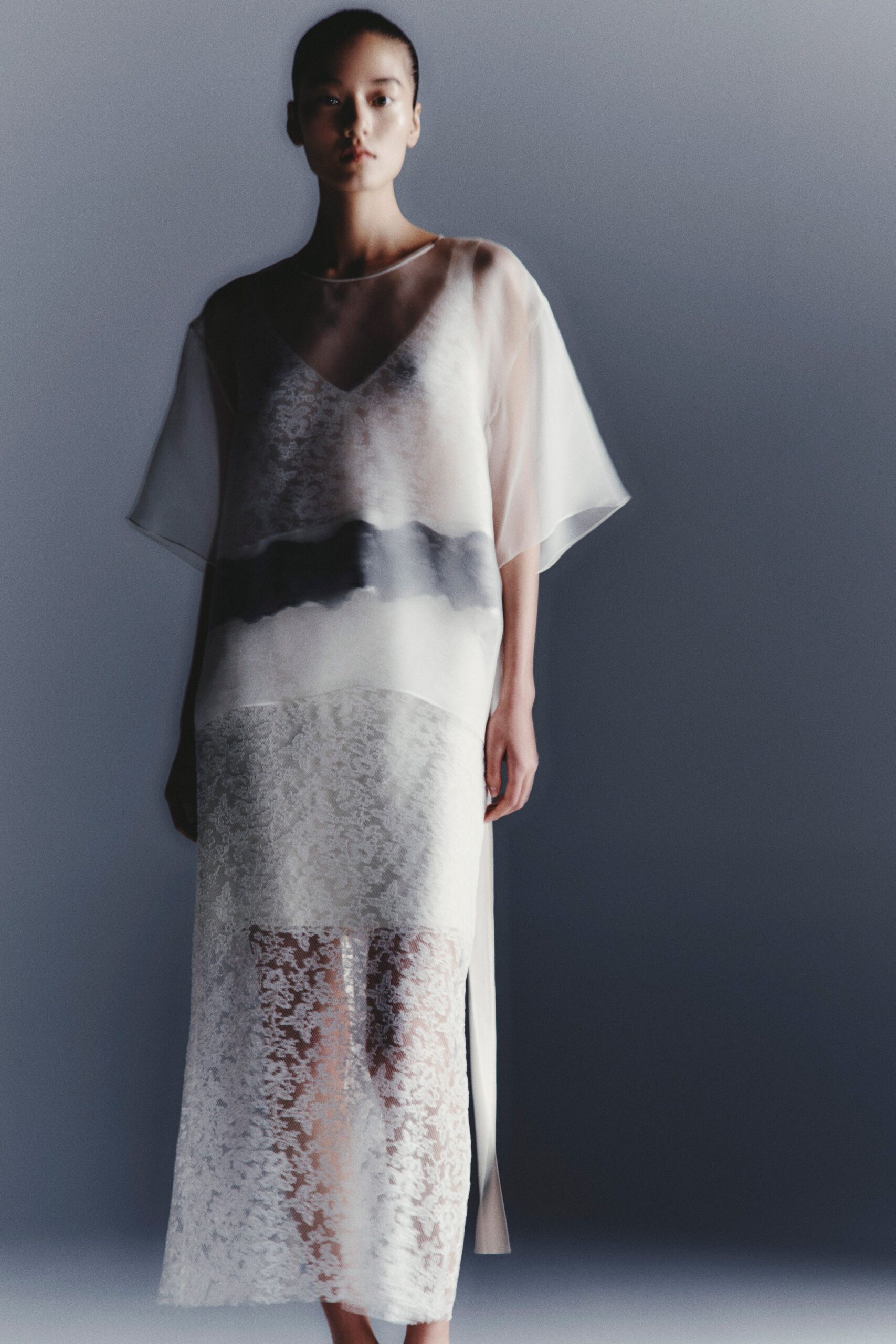 Sheer ivory tunic with a boat neck and inky blurred band. White floral lace slip dress, V neckline ghosting through the chiffon. Long side panel hanging straight past the calf slit. First ferry out, mist still caught on the railings.