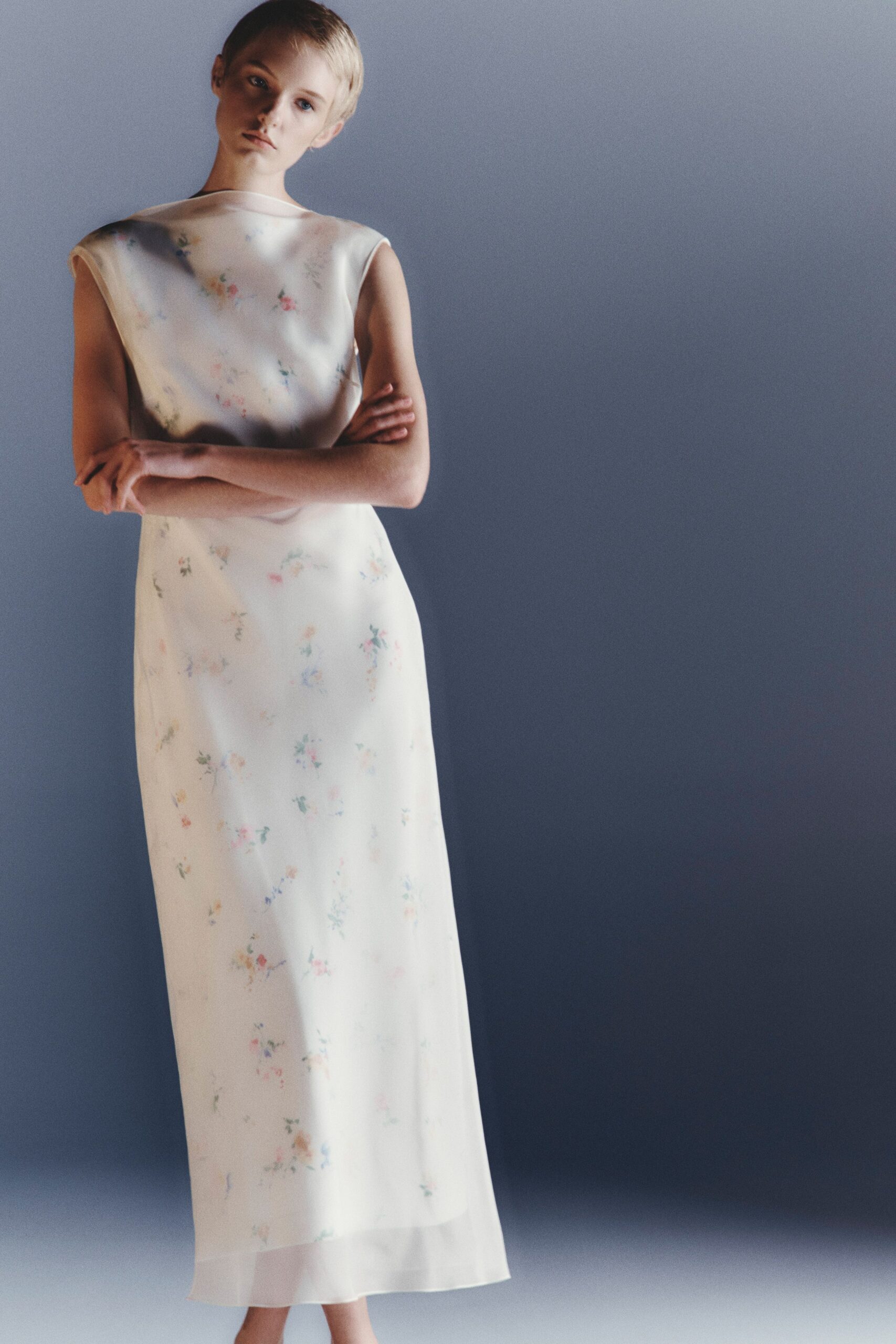 Ivory satin column dress with a gauzy overlayer and washed floral sprigs. Sheer organza band at the ankle, the underlayer stopping short. Sunday reception room light, flowers already starting to drop petals.