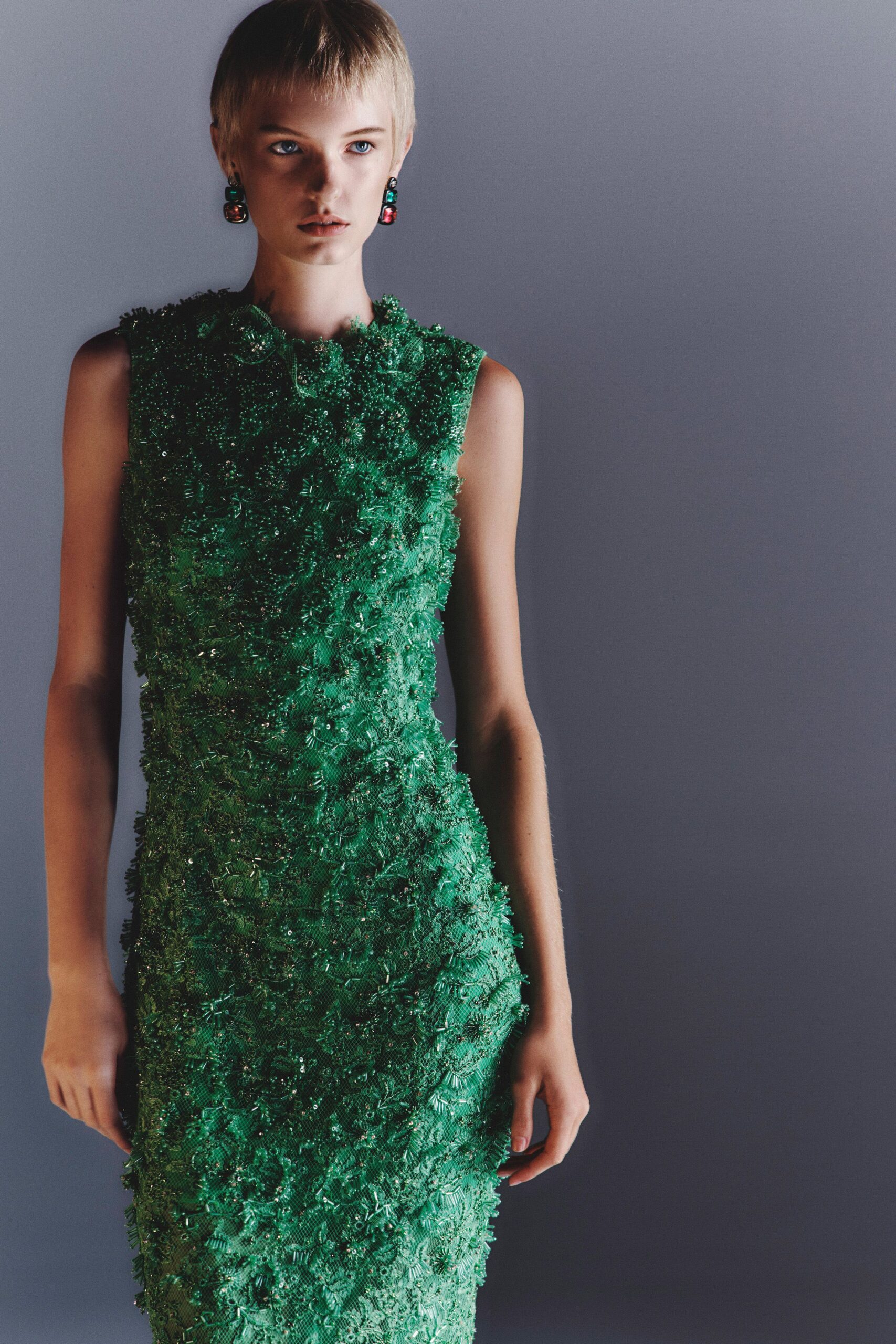 Emerald sleeveless column dress densely crusted with sequins, bugle beads, and raised rosettes. Stacked jewel-drop earrings in emerald and garnet glass. Museum fundraiser, standing under the aquarium glow after speeches end.