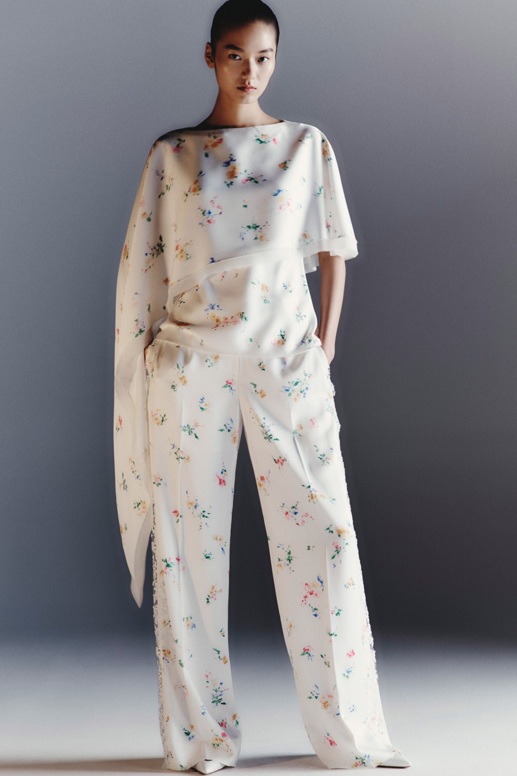 Ivory floral tunic in fluid crepe, one side falling into a scarf-long sleeve. Matching wide-leg trousers with pressed creases and frayed side seams. White pointed shoes barely showing under the hem. Museum atrium at opening hour, florist buckets still sweating by the door.