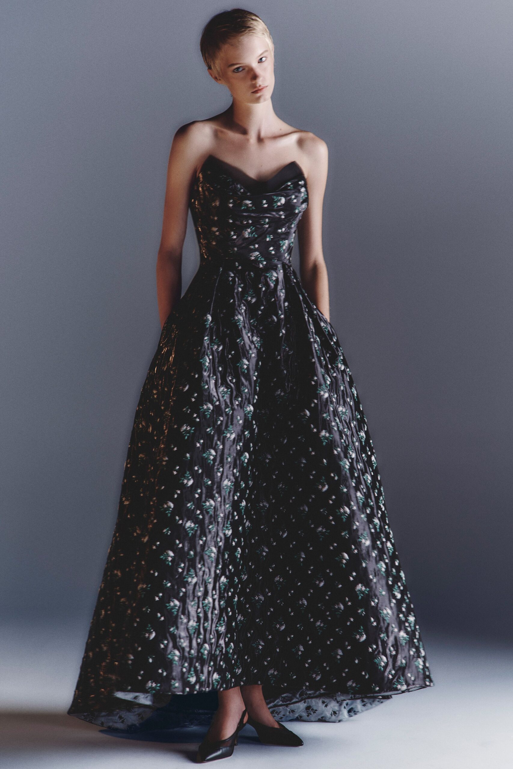 Black strapless ball gown in glossy floral jacquard, sweetheart neckline softly folded. Black pointed flats with a low slingback strap. After the string quartet ends, crossing gravel toward the dark lawn.