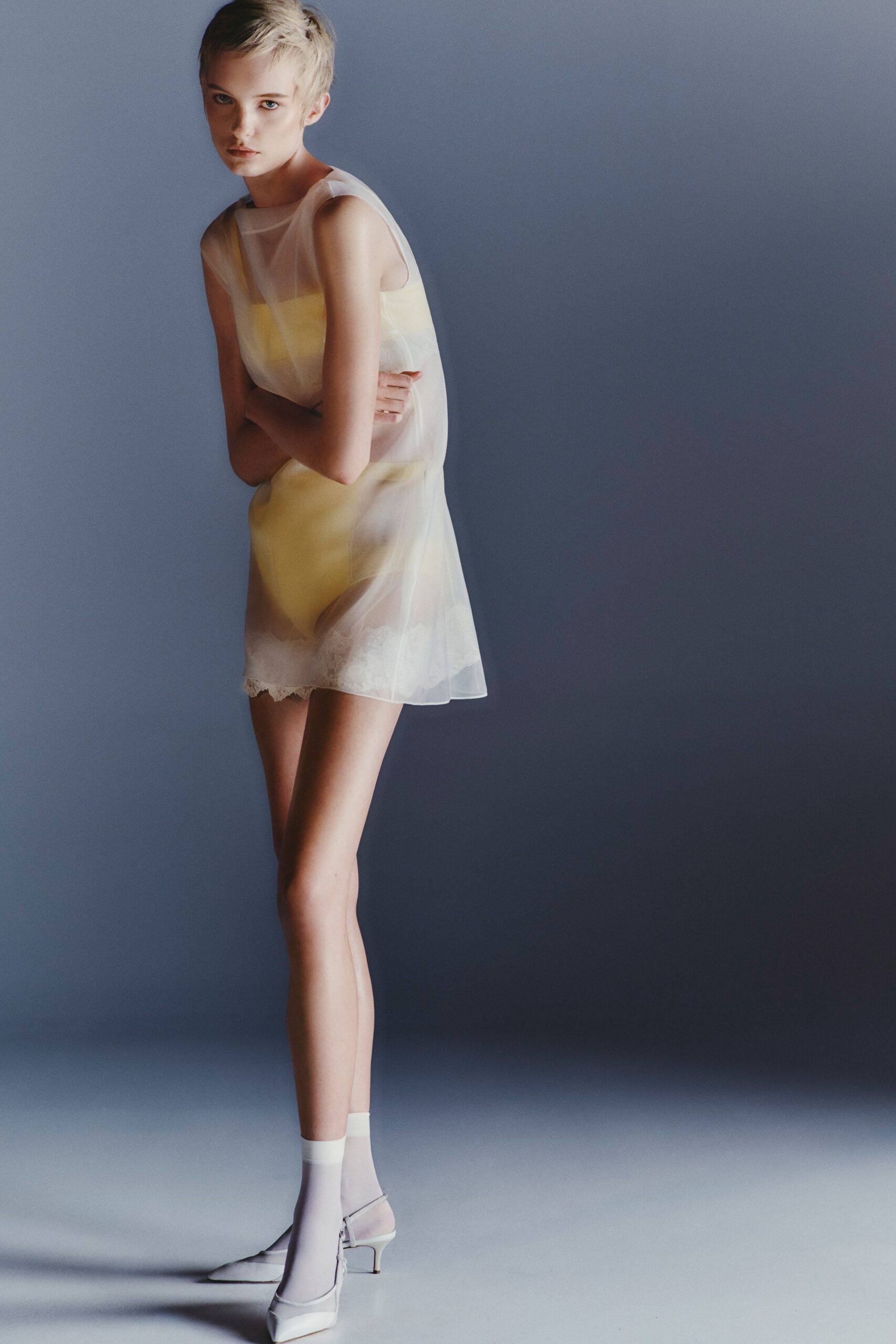 Sheer milky organza shift, the armhole cut wide and square. Butter-yellow bodysuit glowing through the dress. White lace edging peeking along the lower hem. Opaque white ankle socks. White slingbacks with a folded vamp and narrow kitten heel. First light in a hotel room, still dressed before checkout.