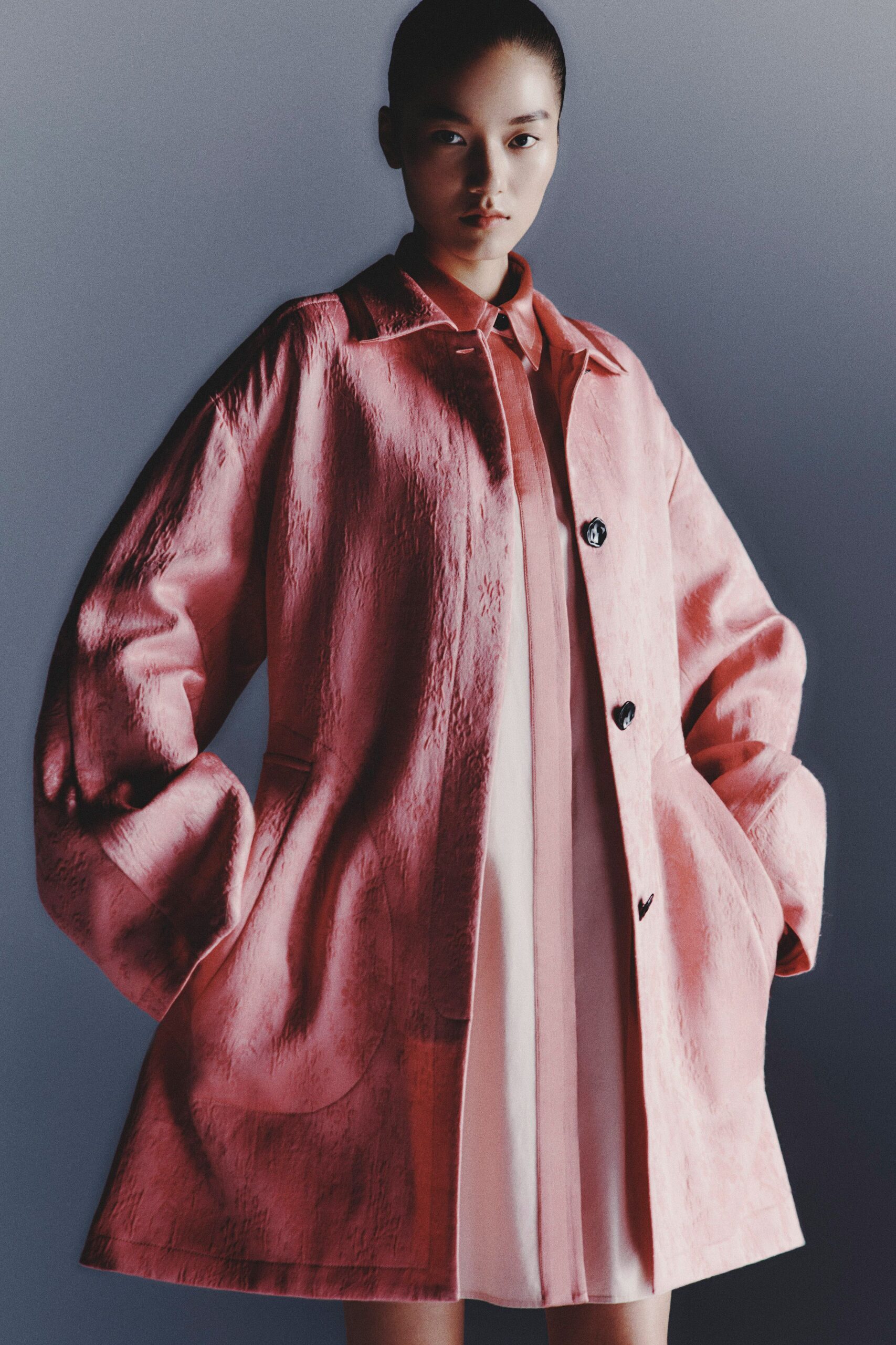 Blush crinkled satin coat with swollen sleeves and black marbled buttons. Pale rose pointed-collar shirt dress peeking below, placket hanging straight. Waiting by the hotel lift before a wedding breakfast turns loud.