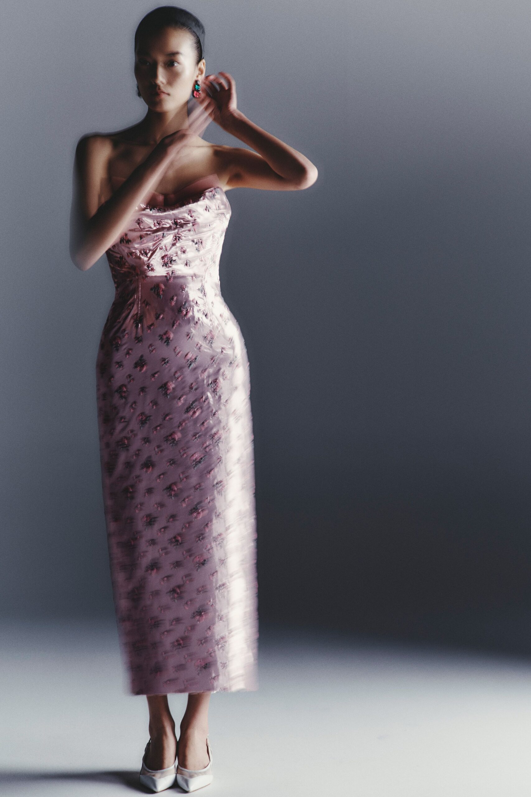 Turquoise-and-raspberry drop earrings catching beside the jaw. Blush strapless column dress in glossy floral jacquard, neckline softly crumpled. White pointed pumps with translucent mesh at the vamp. Waiting by the wedding cake before anyone finds the knife.