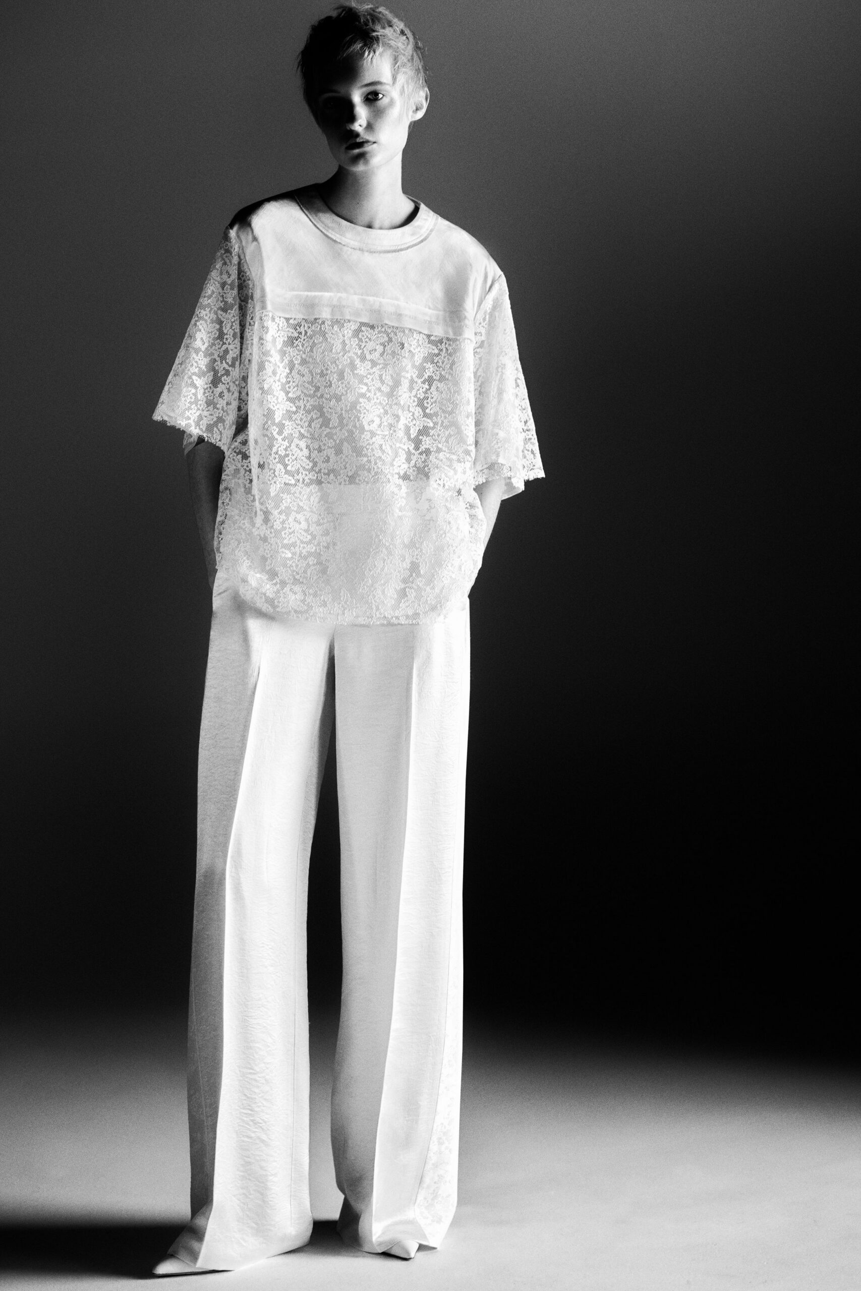 Oversized white lace top with a satin yoke and dropped sleeves. White wide-leg trousers in fluid crepe, pooling lightly over the shoe. Sharp white points just peeking from the hem. Morning after the wedding, hotel curtains still half closed.