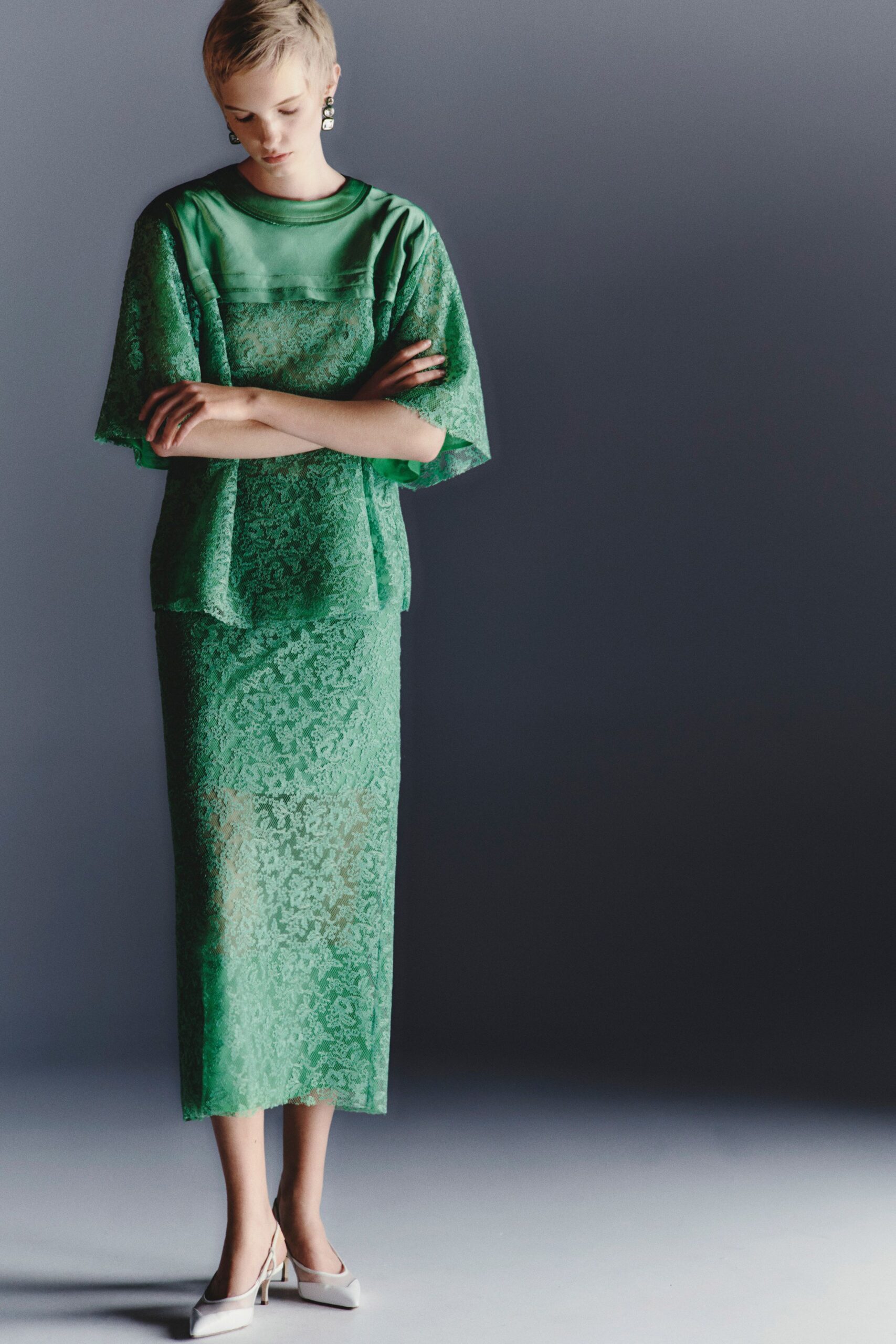 Stacked square drop earrings in darkened silver and milky stones. Emerald lace top with a satin yoke and wide elbow sleeves. Matching straight midi skirt, lace laid over a pale underlayer. White leather mules with a narrow flared heel. Hotel corridor before breakfast, still wearing last night's earrings.