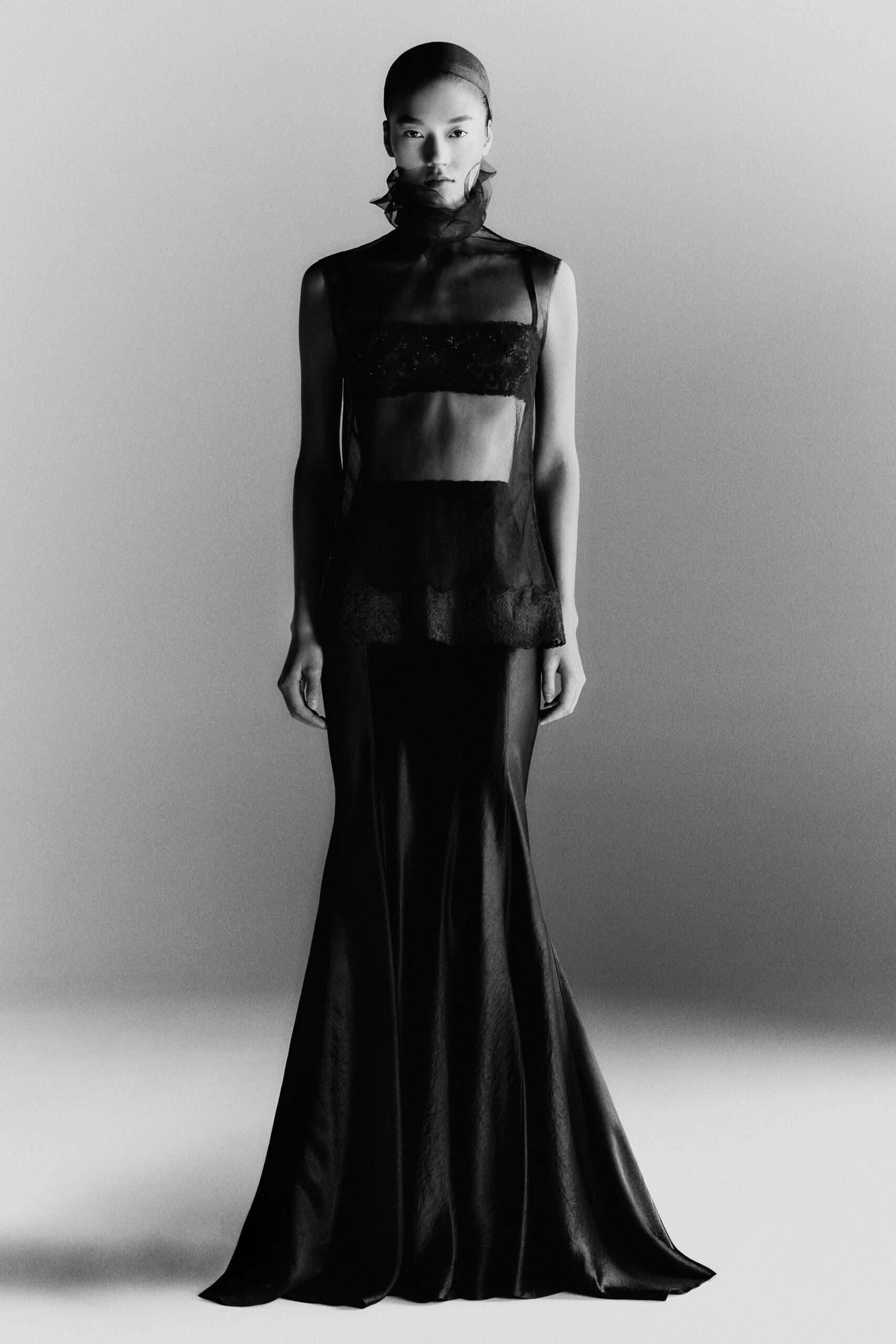 Sheer black organza shell with a high ruffled neck. Sequined bandeau catching light beneath the transparent layer. Black satin column skirt pooling softly at the floor. Museum fundraiser, standing beside the darkened fountain after midnight.