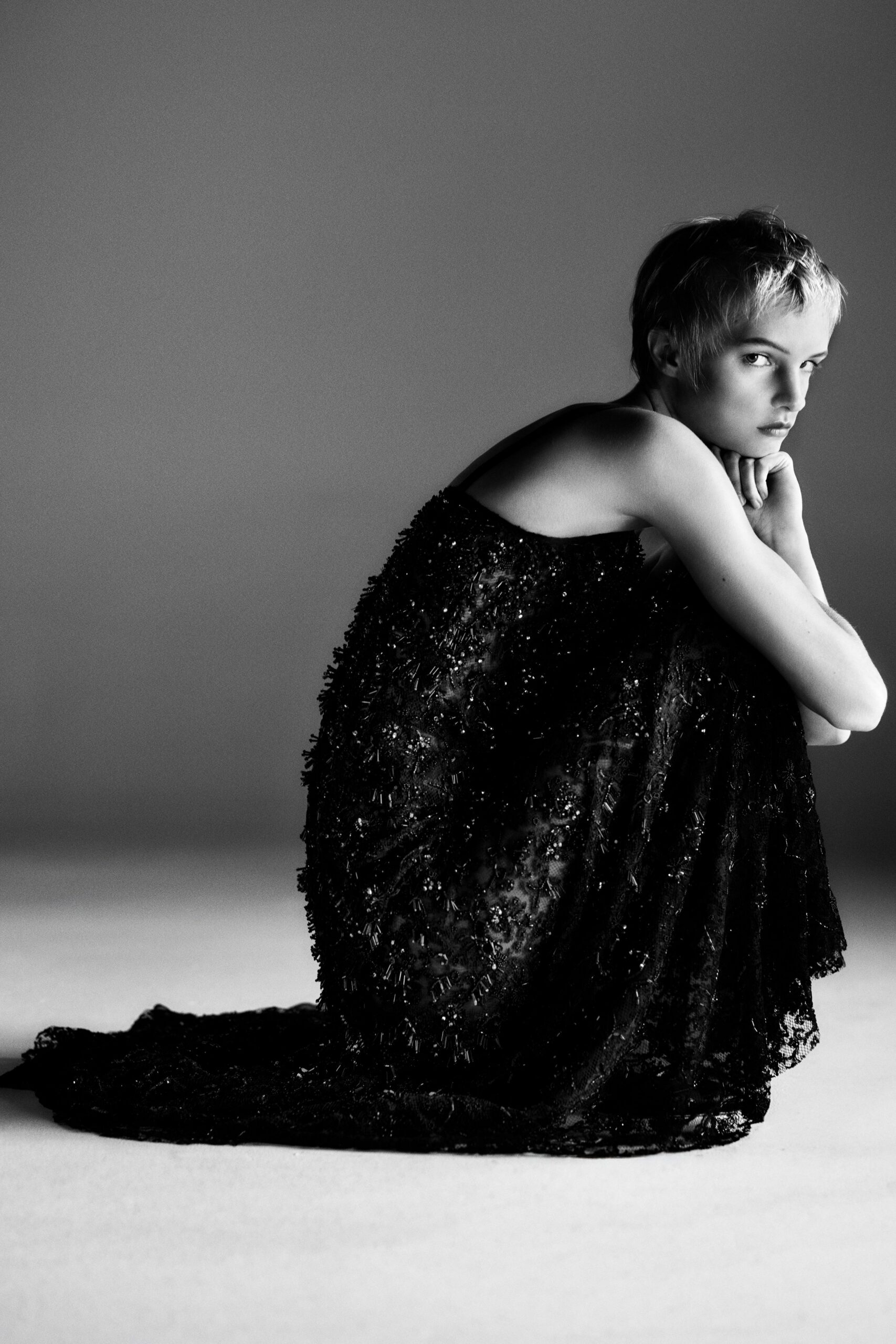 Black lace gown crusted with bugle beads and shaggy fringe. Long scalloped lace train pooling flat behind her knees. Morning after the hotel ball, curtains still closed against the sea.