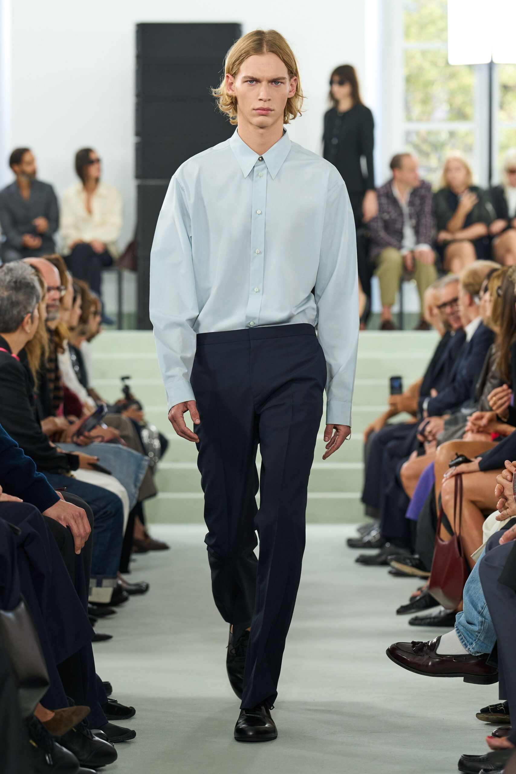 Pale blue cotton shirt with long cuffs and a pinched spread collar. Dark wool trousers cut straight, crease running clean to the shoe. Black leather derbies with a blunt rounded toe. For the first meeting after summer when nobody mentions the tan.