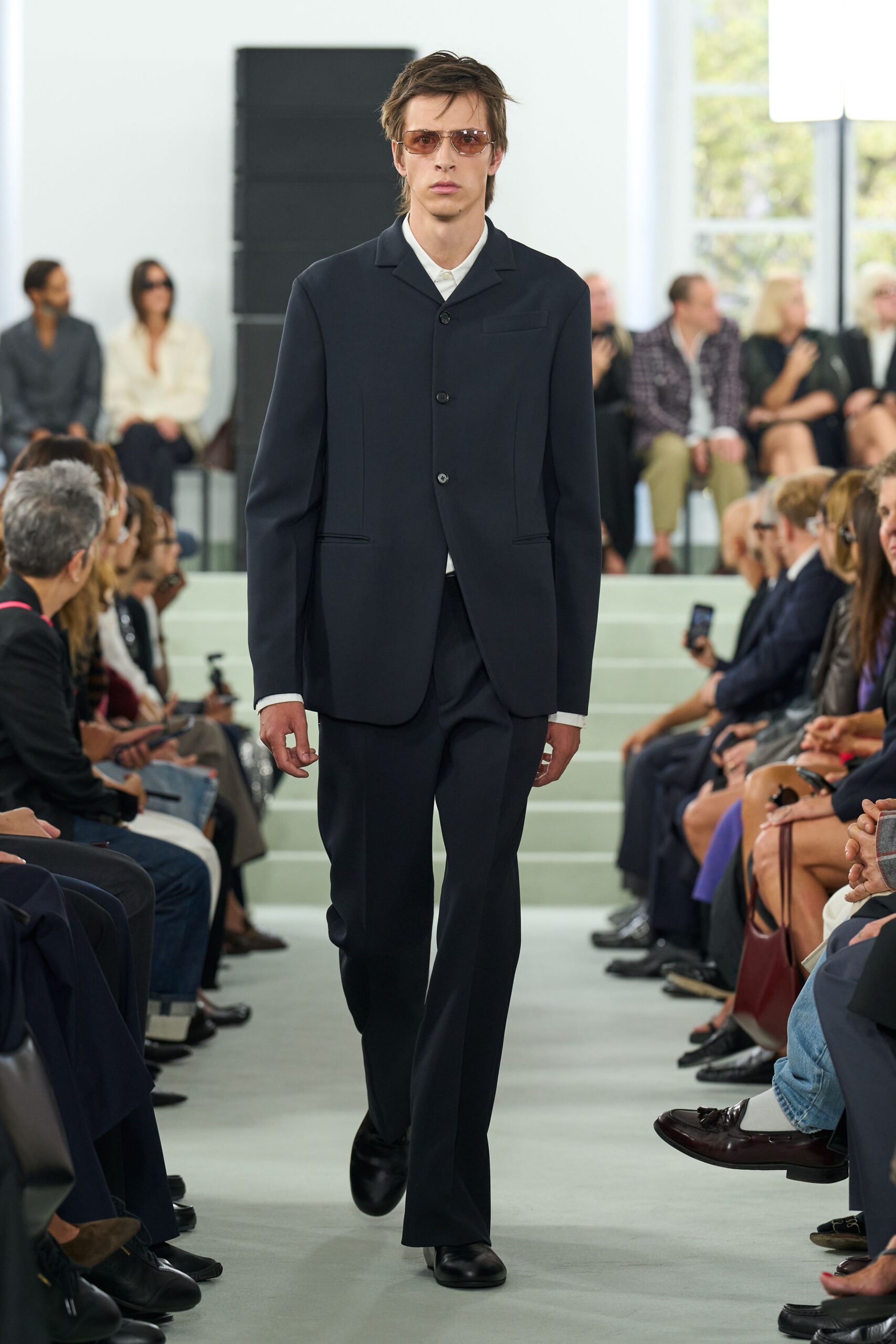Amber-tinted aviator sunglasses with a thin metal bridge. Inky wool jacket with a low stance and flapless welt pockets. White shirt buttoned clean to the throat. Matching straight trousers breaking softly over the shoe. Black leather derbies, rounded and mirror-shined. Crossing a quiet municipal atrium before the offices open.