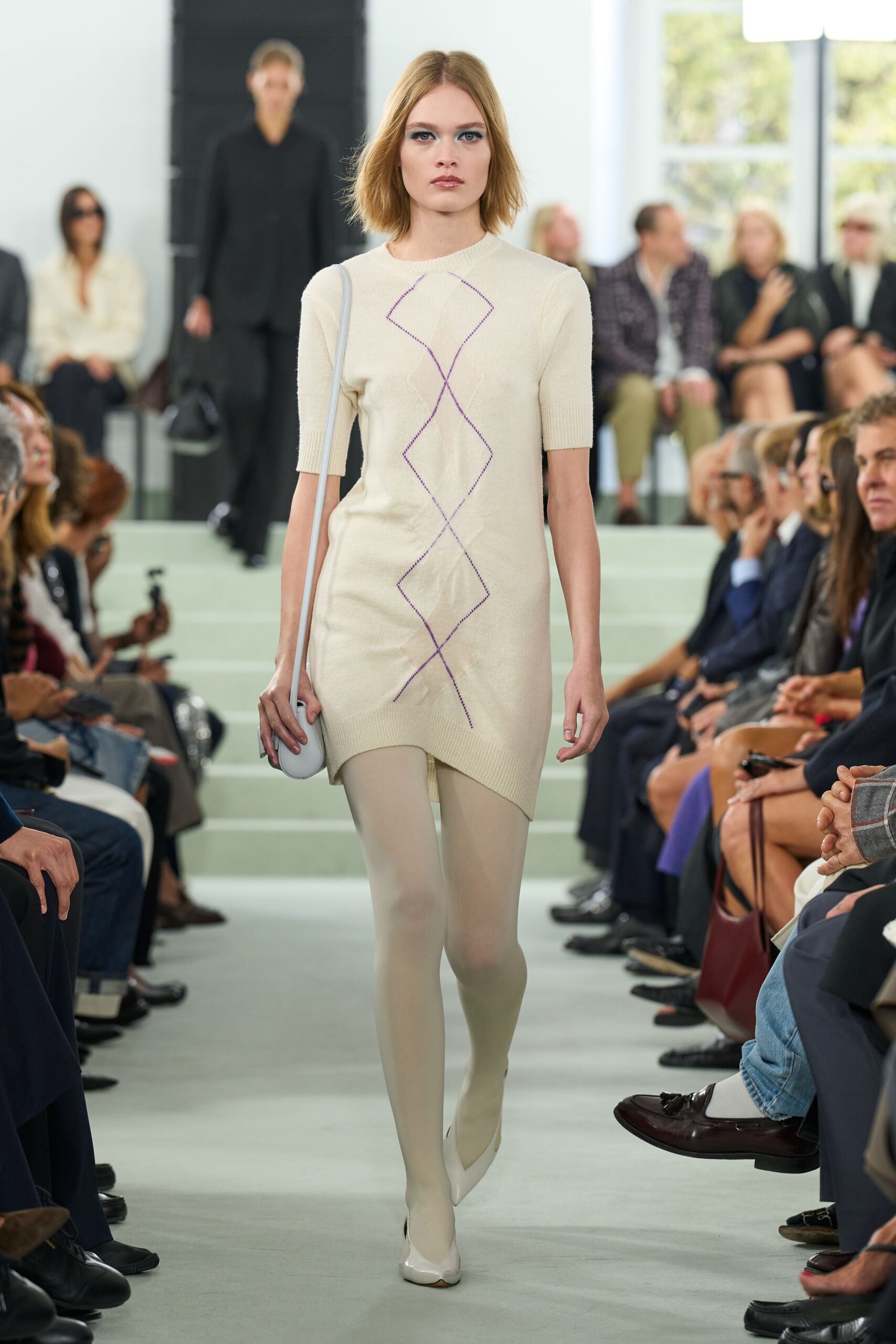 Cream fine-gauge knit dress with violet argyle lines and an uneven hem. Opaque ivory tights. Pale grey oval bag on a skinny strap. Milk-white pumps with a split, petal-like vamp. Walking into a quiet gallery atrium while the morning is still cool.