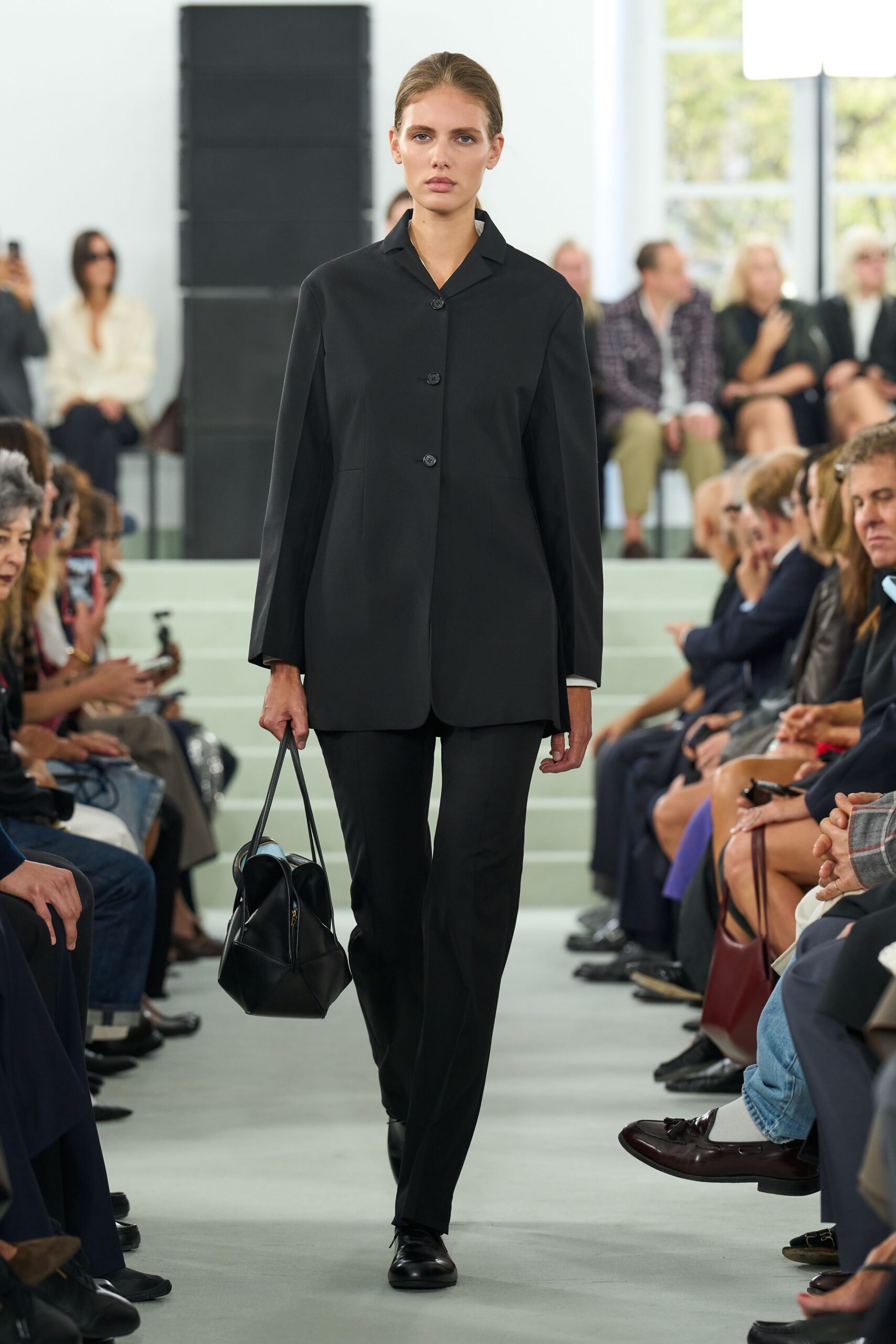 Long black jacket in compact suiting, three small buttons and rounded hems. Straight black trousers with a sharp front crease. Black leather holdall, folded into soft geometric panels. Black leather flats with squared toes. For crossing a quiet museum atrium before the doors open.