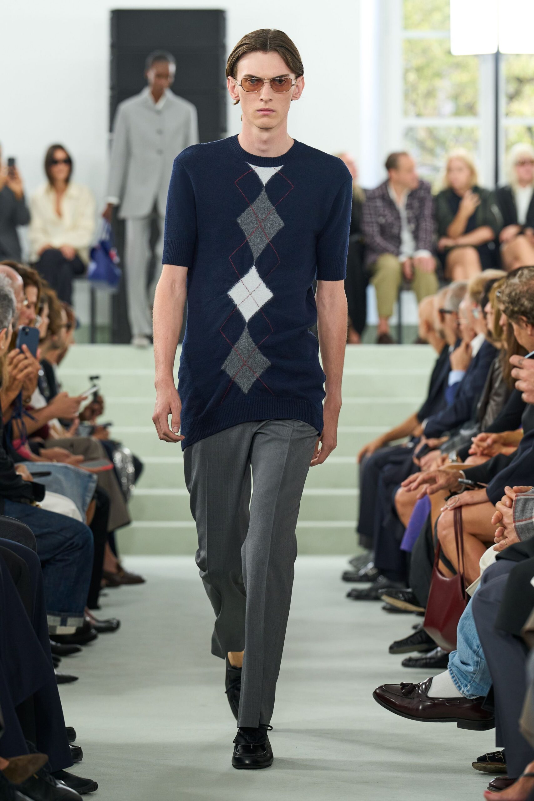 Amber-tinted aviator sunglasses with a thin gold bridge. Navy short-sleeve knit sweater, elongated hem, argyle column drifting off center. Soft grey tailored trousers with a pressed crease breaking at the shoe. Black square-toe loafers. Walking into a quiet members' club before lunch service starts.