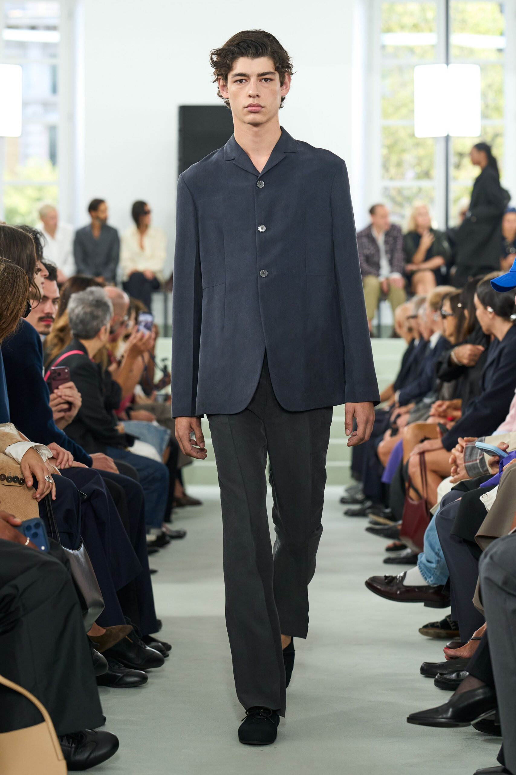 Ink-blue tailored jacket with a soft camp collar and three horn buttons. Charcoal straight trousers with a long break pooling slightly at the shoe. Black suede lace-ups, blunt and almost slipper-flat. Crossing a hushed museum atrium before the security gates unlock.
