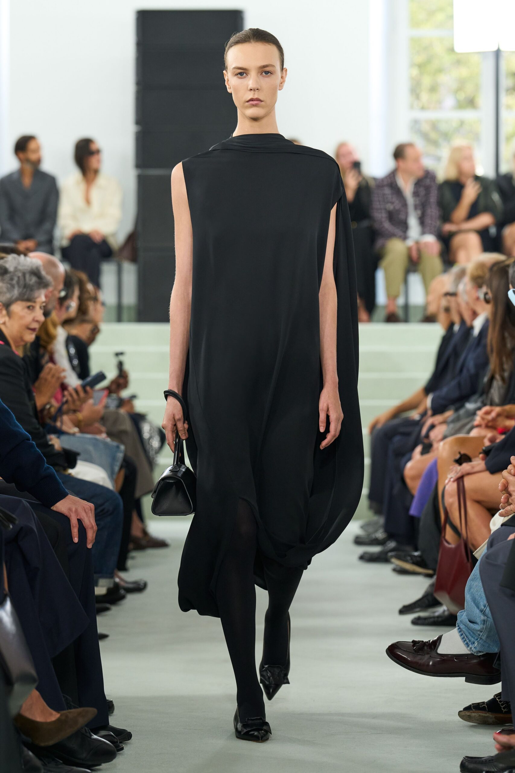 Black jersey column dress with a draped cowl neck and cape side panel. Opaque black tights. Thick black bangle. Small black top-handle bag with a rounded gusset. Pointed black flats with a narrow strap across the vamp. Crossing a silent museum atrium before the doors open to the public.