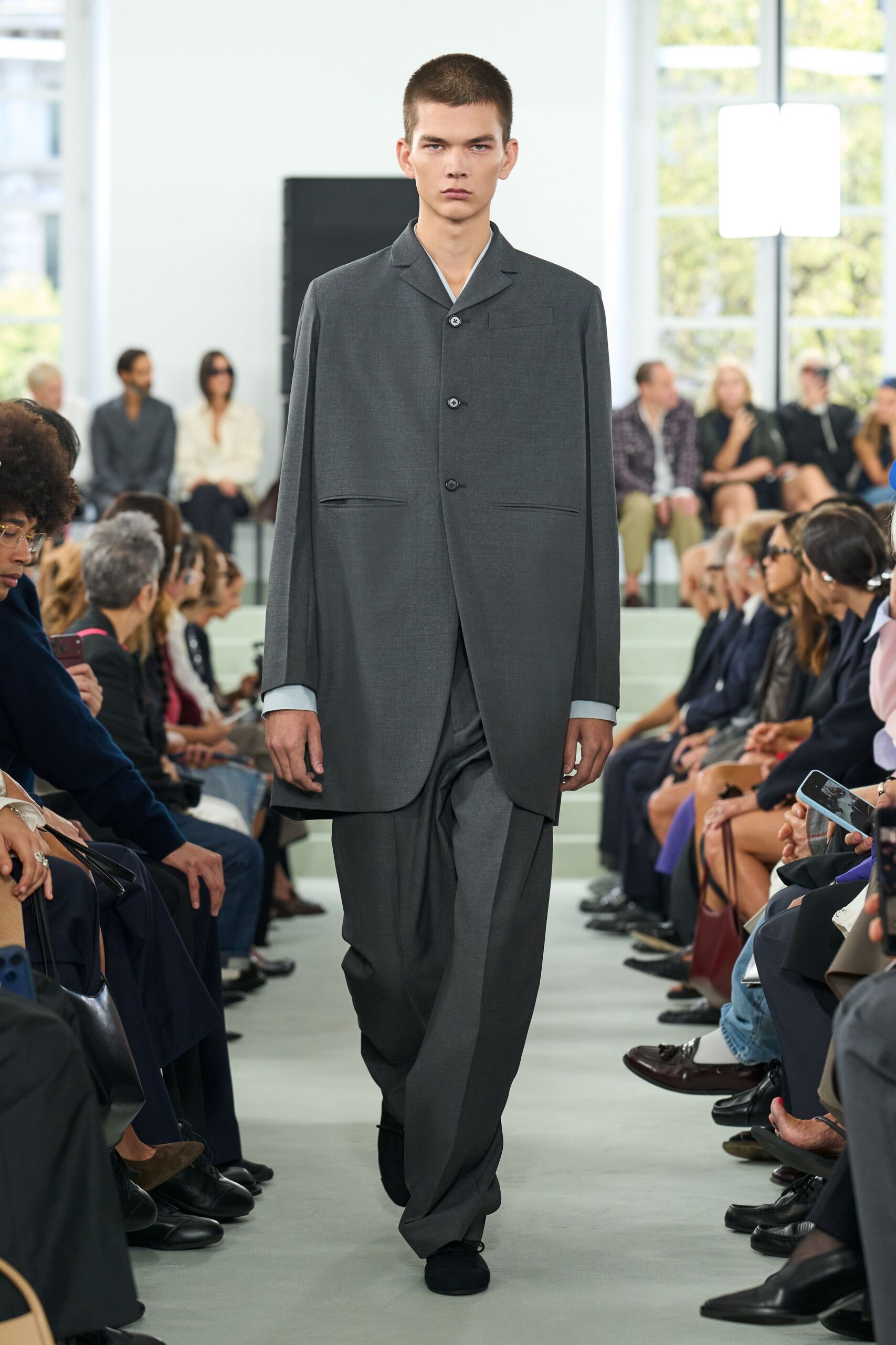 Charcoal worsted coat cut long and clean, with slashed welt pockets. Pale blue cuffs and V-neck peeking sharp against the lapel. Matching wide trousers breaking deep over the shoe. Black suede shoes, almost swallowed by the hem. Crossing a quiet civic atrium before offices open upstairs.