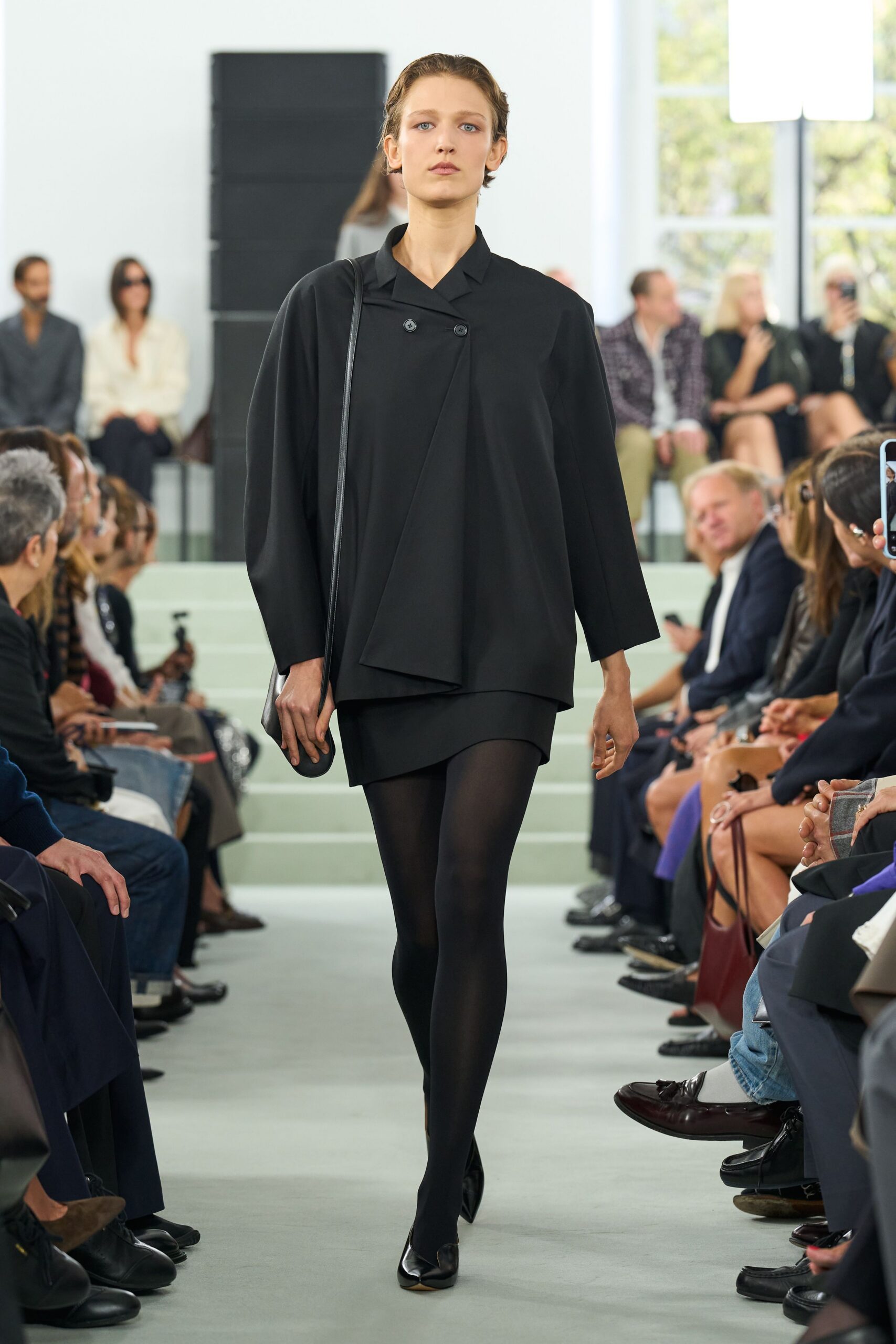 Black crepe tunic jacket with offset double buttons and a folded front panel. Short black skirt curving lower at one hip. Opaque black tights. Slim black leather strap and small pouch tucked at the hip. Pointed black flats with a pale split sole peeking through. For crossing a hushed museum atrium before the doors open.