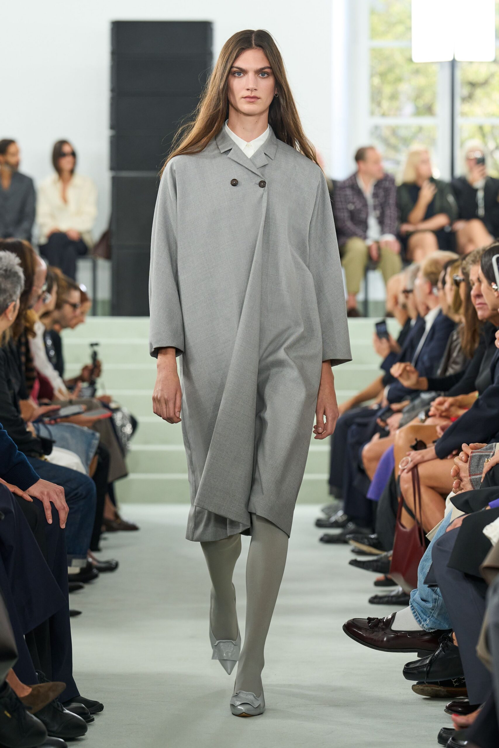 White point-collar shirt buttoned tight at the throat. Pale grey wool coat dress, double-breasted with a long off-center drape. Matching grey opaque tights. Pointed grey pumps with a scalloped vinyl cuff. Crossing a quiet municipal atrium before the offices open.