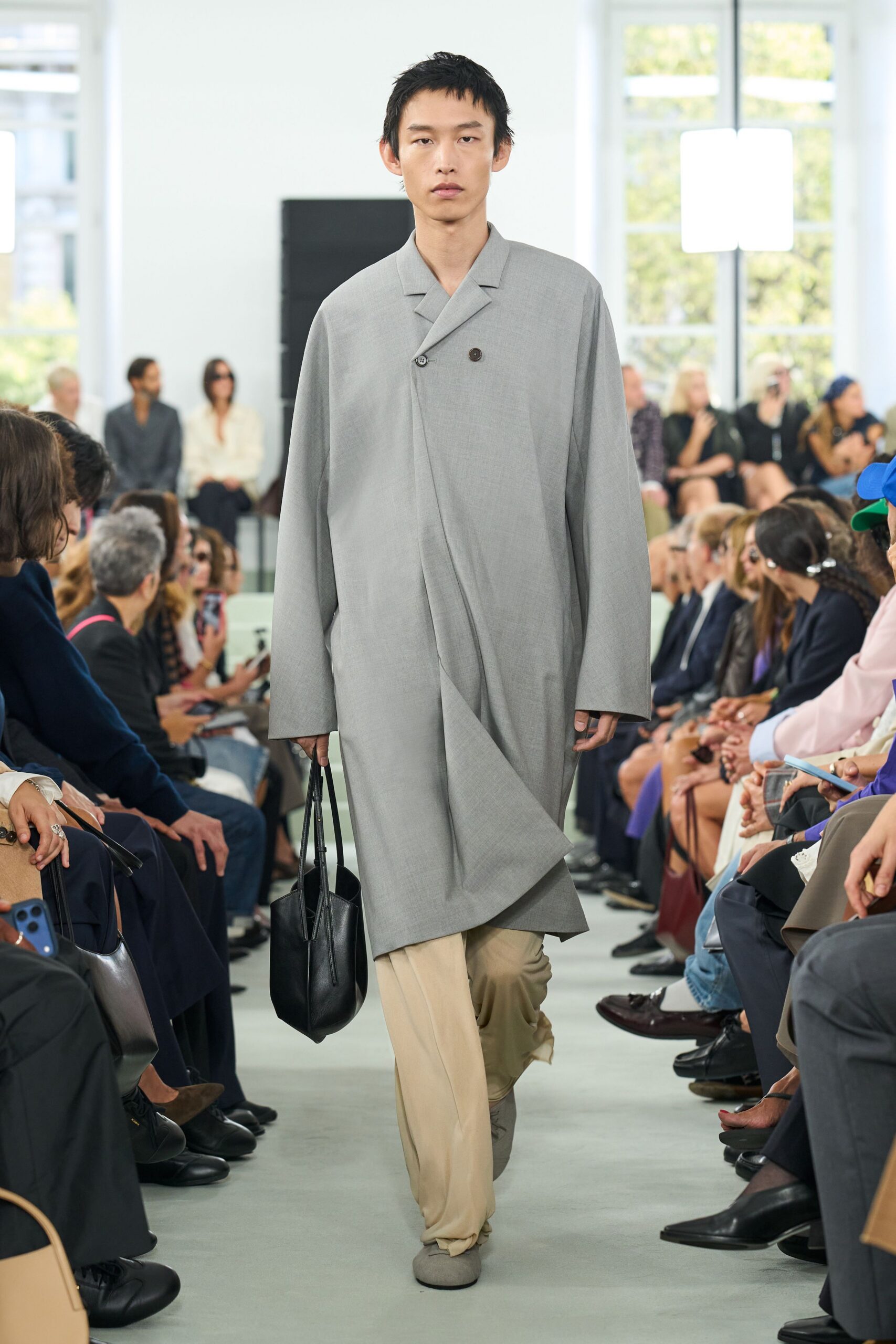 Pale grey wool coat cut with a sweeping wrapped front panel. Cream trousers puddling softly over the instep. Black leather bucket bag with long narrow handles. Ash suede slip-ons, blunt at the toe. Crossing a quiet museum atrium before the doors open to the public.