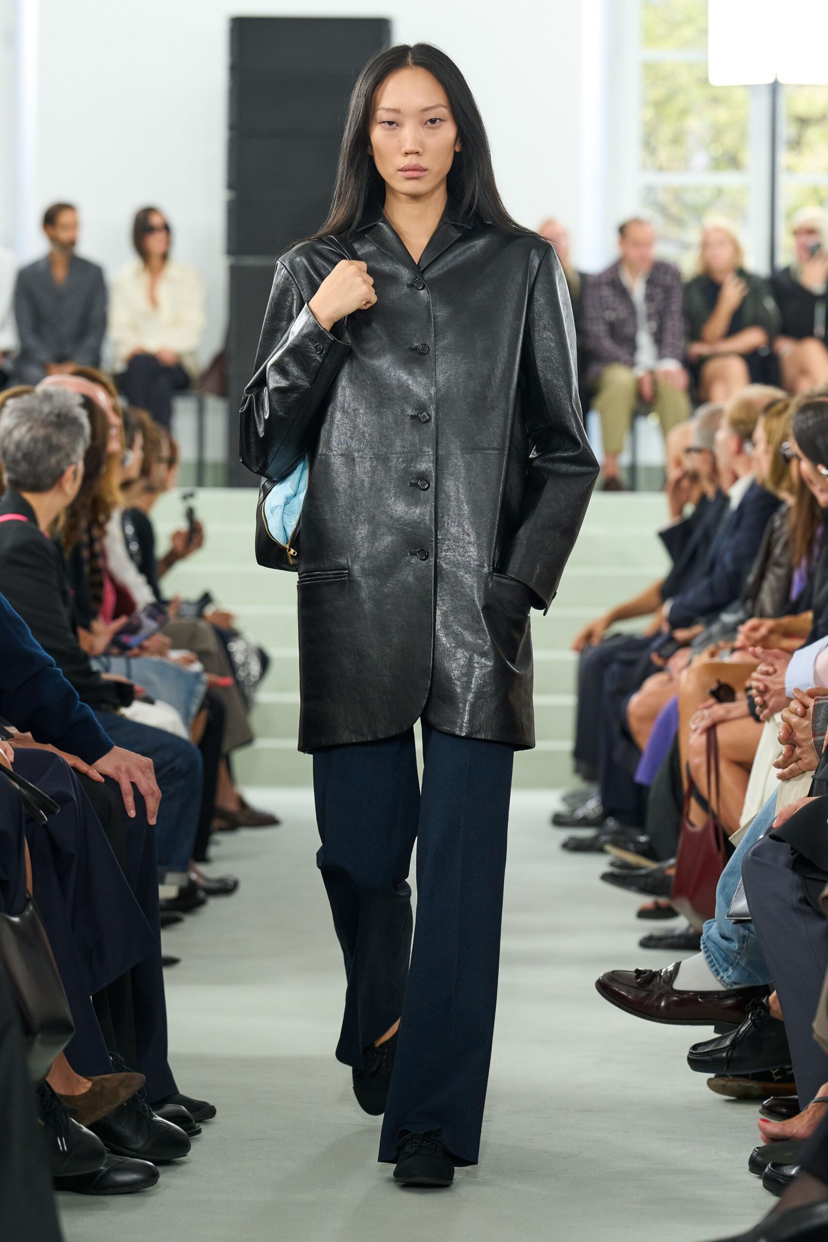 Black leather coat with a dry sheen, long and slightly boxy. Deep navy trousers breaking softly over the shoe. Black lace-up flats, almost swallowed by the hem. Powder-blue lining flashing from a half-open black leather bag. For crossing a white lobby before anyone has had their second coffee.
