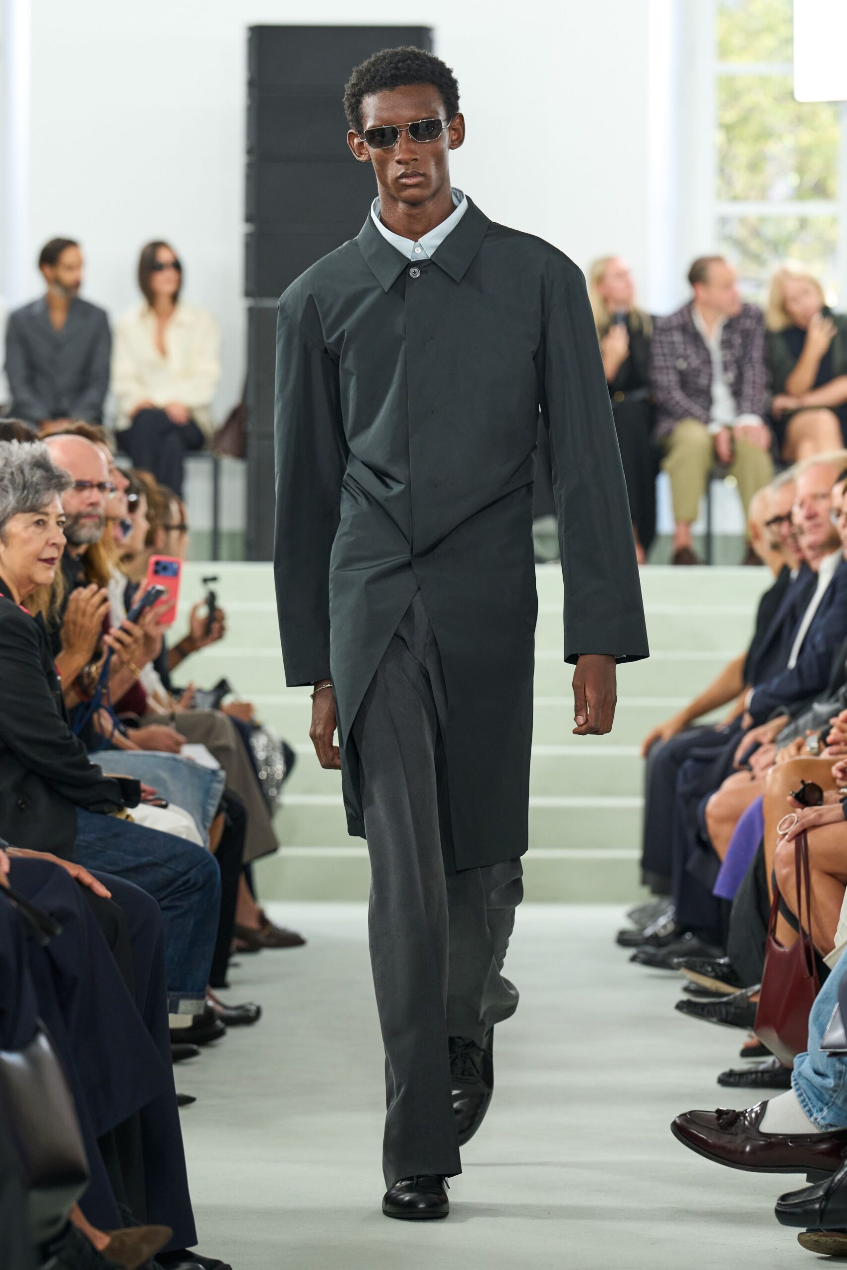 Narrow black-lensed sunglasses with thin silver wire rims. Pale blue shirt buttoned clean to the throat. Ink-dark coat with a concealed placket and skewed wrapped front. Charcoal trousers falling in a soft, wide straight leg. Black leather lace-ups with rounded toes. For crossing a glass office atrium before anyone else is in.