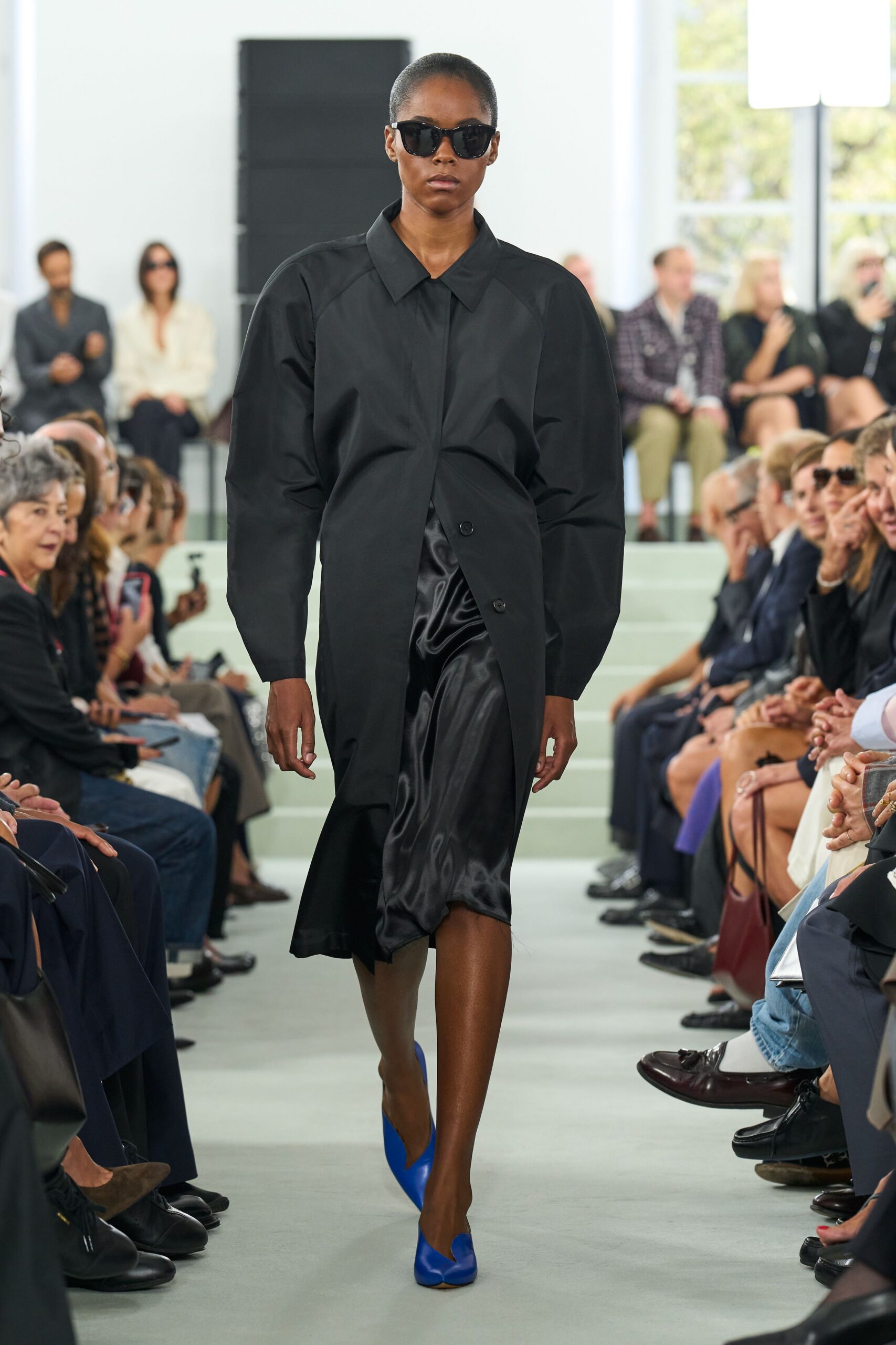 Glossy black cat-eye sunglasses. Black coat with puffed raglan sleeves and a slightly twisted button placket. Black satin slip skirt, liquid shine pooling into a skewed hem. Cobalt leather pumps with split vamp openings. Crossing a museum atrium before the rain starts properly.