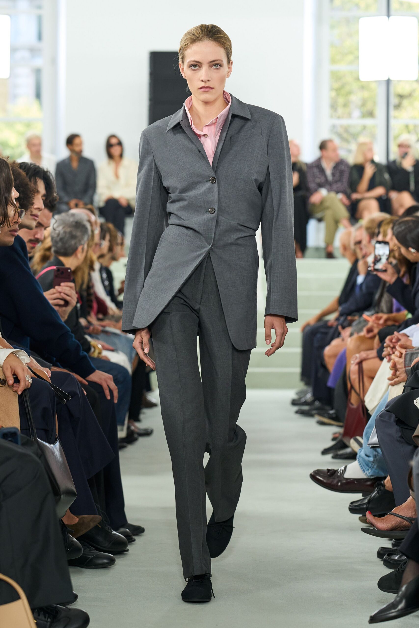 Pale pink shirt with a soft crumpled collar peeking above the lapel. Charcoal suiting jacket cut long, twisted shut with a low third button. Matching straight trousers with a sharp crease breaking over the shoe. Black lace-up shoes in matte suede. For the first meeting after lunch when the room still smells of coffee.