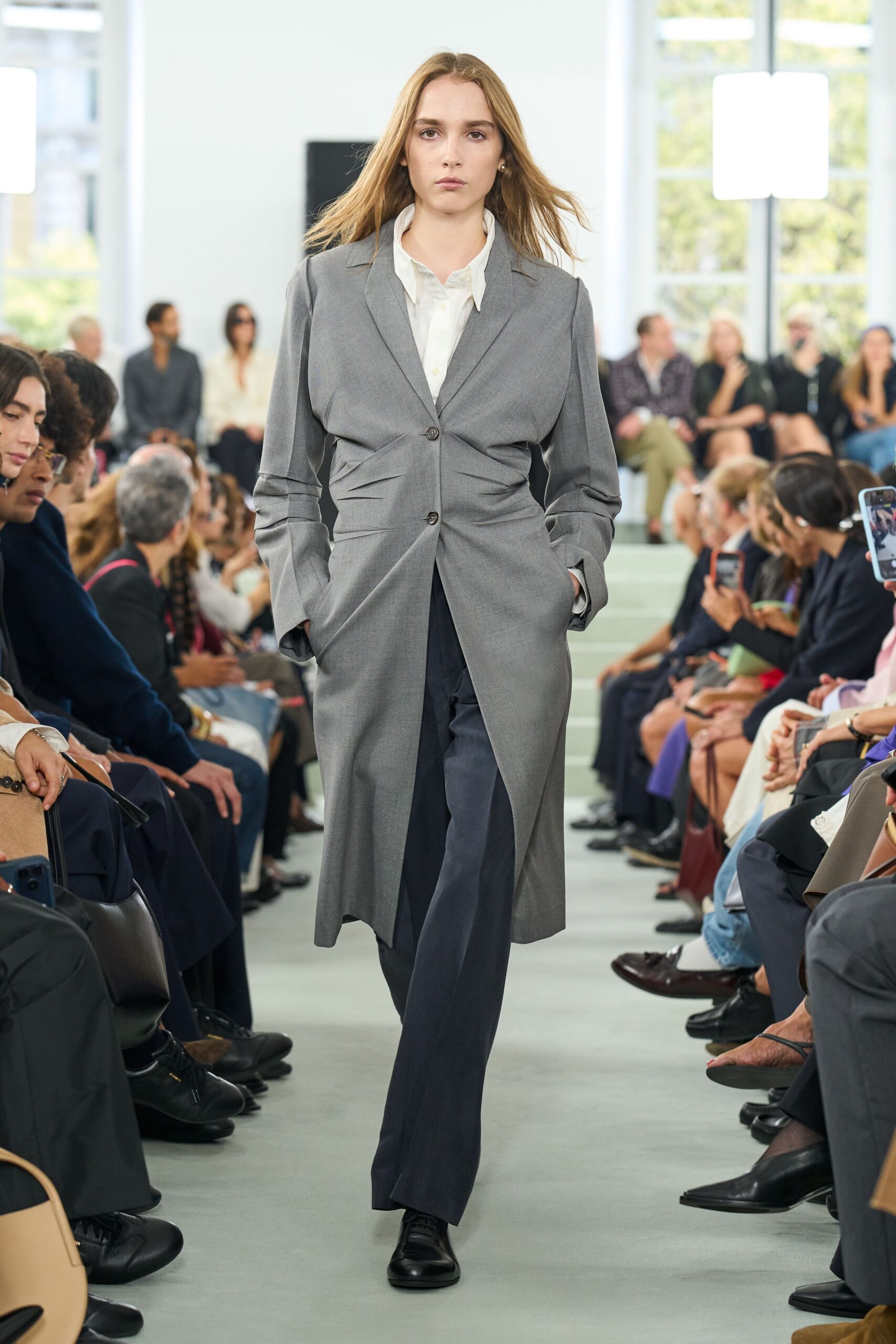 Long dove-grey tailoring coat, pinched with folded tucks around the waist. Soft white shirt, collar points lifting loose from the neck. Dark straight trousers breaking lightly over the instep. Black leather lace-ups, plain rounded toes. For the quiet hour before a courthouse wedding, coffee going cold.
