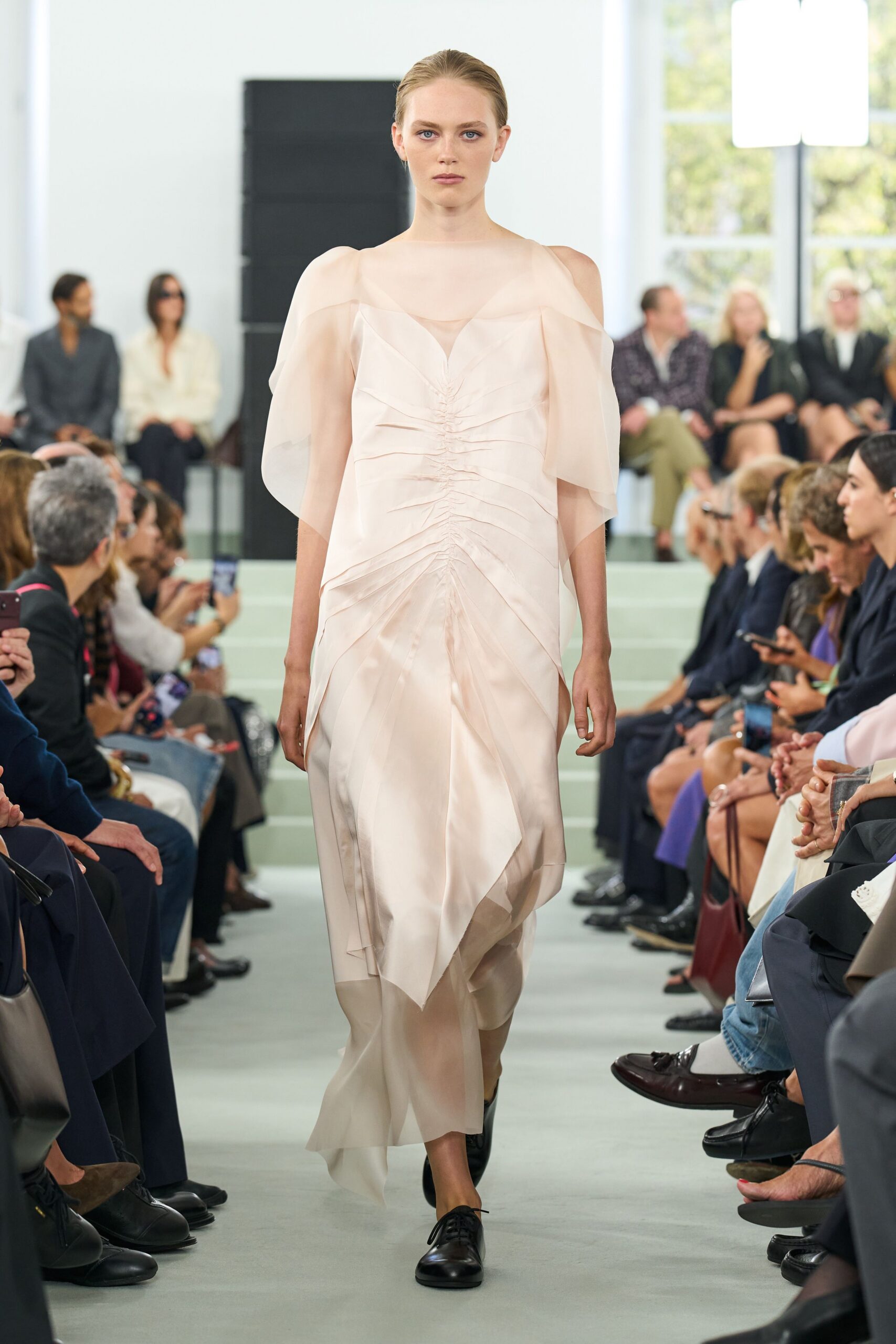 Blush silk-organza dress, ruched straight down the center with floating shoulder panels. Black leather derbies with plain laces under the trailing hem. Crossing a museum atrium before the rain starts again.