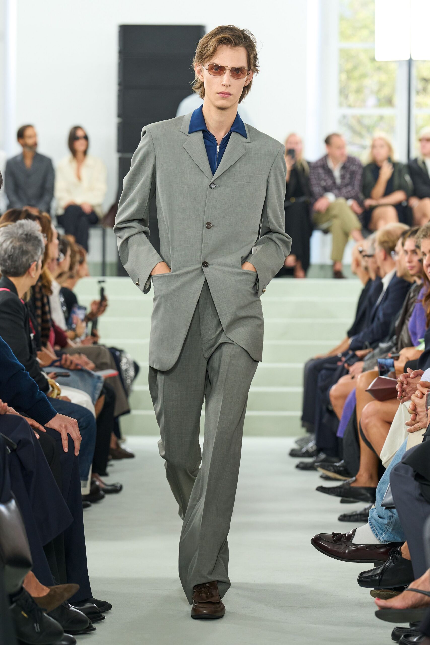 Amber-tinted aviators with a thin silver bridge. Pale grey suiting blazer, long line with low flapless pockets. Cobalt zip polo, wide collar spread over the lapels. Matching straight trousers with a soft break over the shoe. Dark brown square-toe loafers. For crossing a hotel lobby before the conference coffee has cooled.
