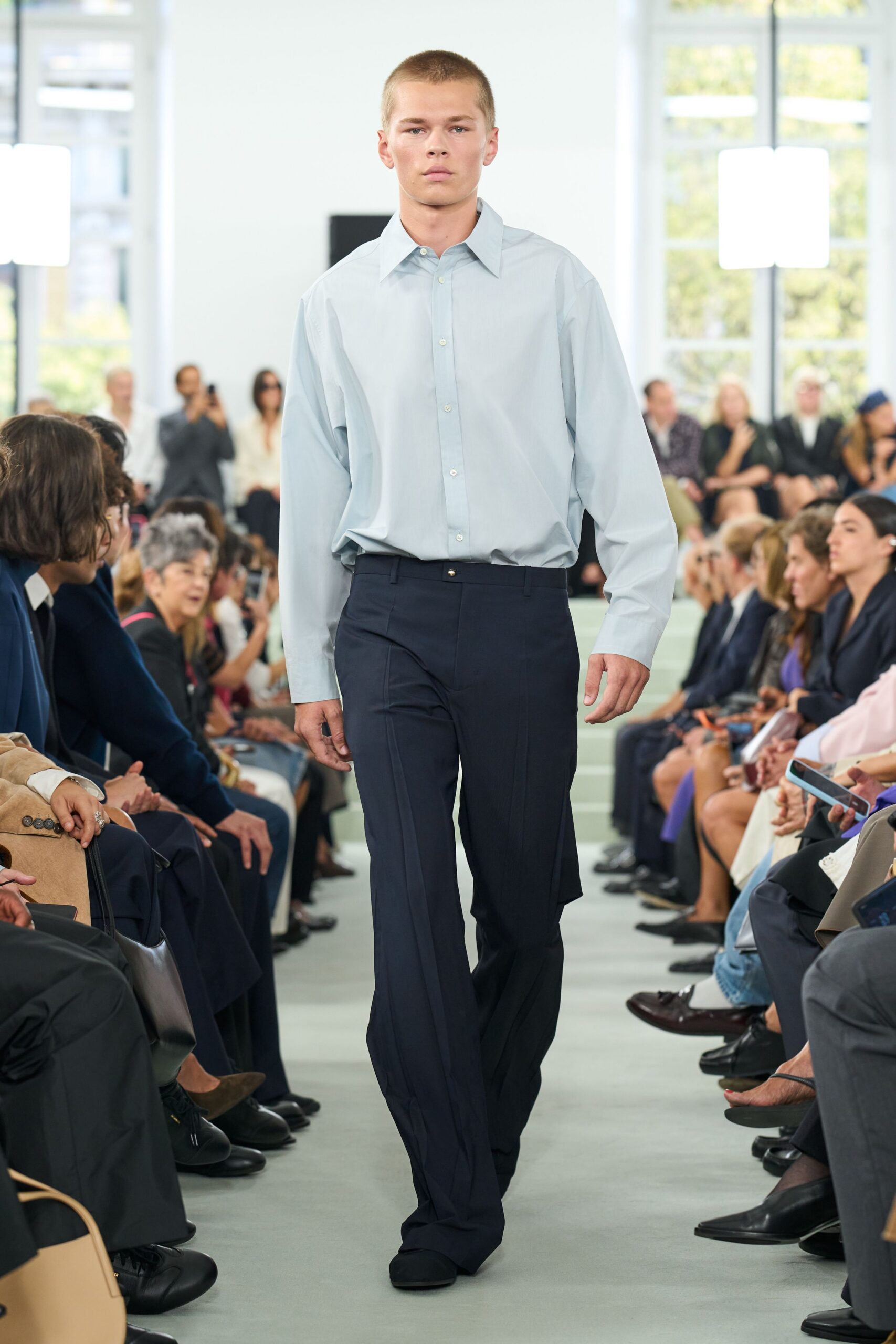 Pale blue cotton shirt, oversized point collar and soft bloused tuck. Deep navy trousers with pressed creases and a single gold waistband stud. Black square-toe shoes disappearing under the break. Early meeting in a glass office, before anyone opens the blinds.