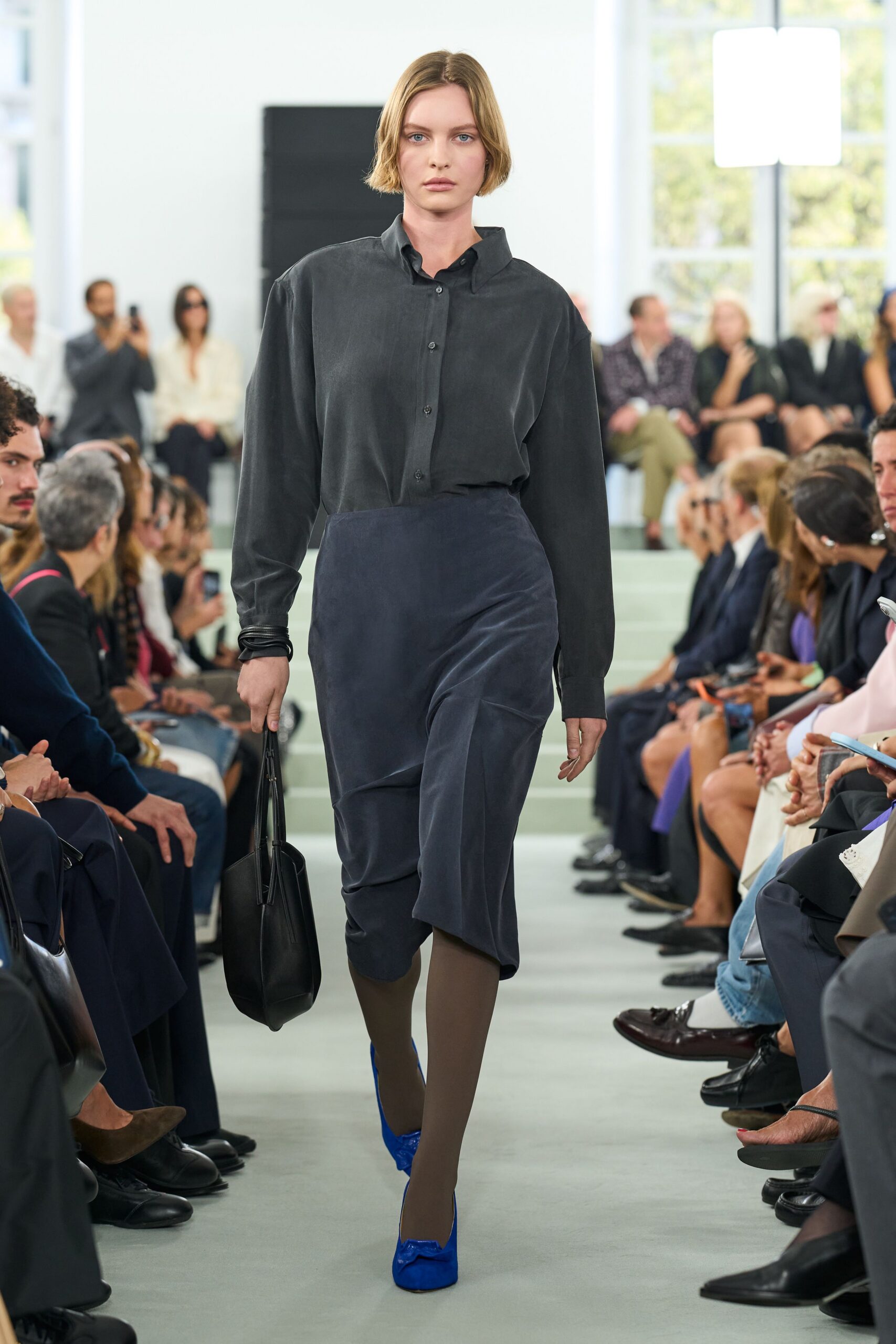 Charcoal silk shirt, one side of the collar kicked upward. Ink draped skirt in velvety suiting, folding across the knees. Stacked black cuffs. Sheer taupe tights. Cobalt satin pumps with blunt square toes. Black leather tote, long handles hanging straight. For the Monday presentation where nobody agrees on the numbers.