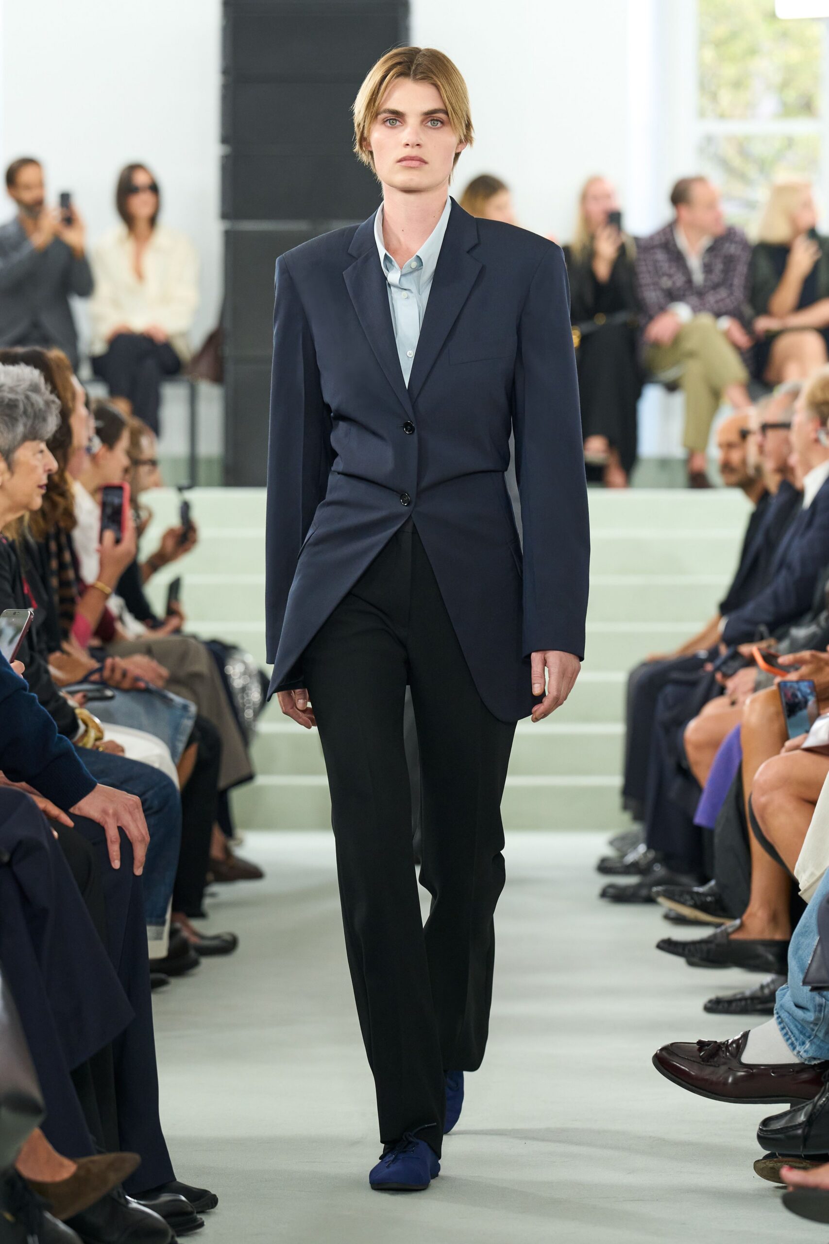 Pale blue shirt in crisp poplin, top button left open. Navy tailored blazer, pinched through the waist with a skewed hem. Black straight trousers with a clean break over the shoe. Cobalt suede lace-ups with a squared-off toe. First meeting after the merger, coffee cooling untouched by your elbow.