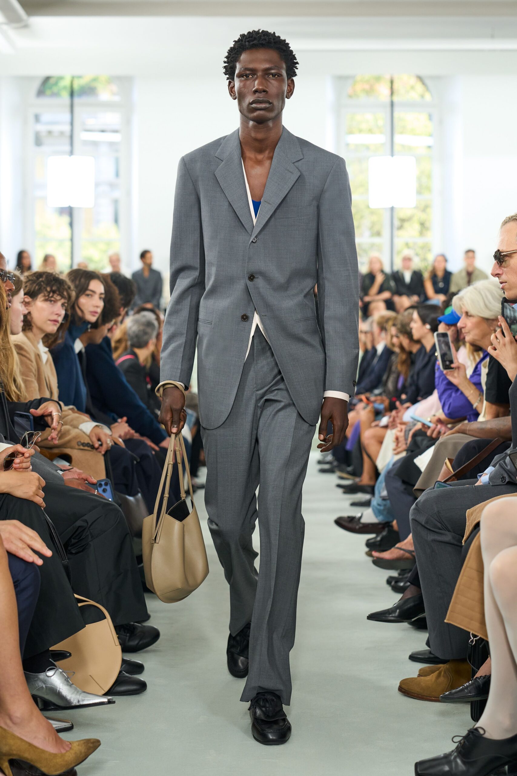 Grey wool blazer with narrow shoulders and a low three-button stance. Cobalt inset peeking from a deep cream V. Matching straight trousers with a pressed front crease. Taupe leather bucket bag with long doubled handles. Black square-toe shoes. Walking into a museum board meeting with a beach bag by mistake.