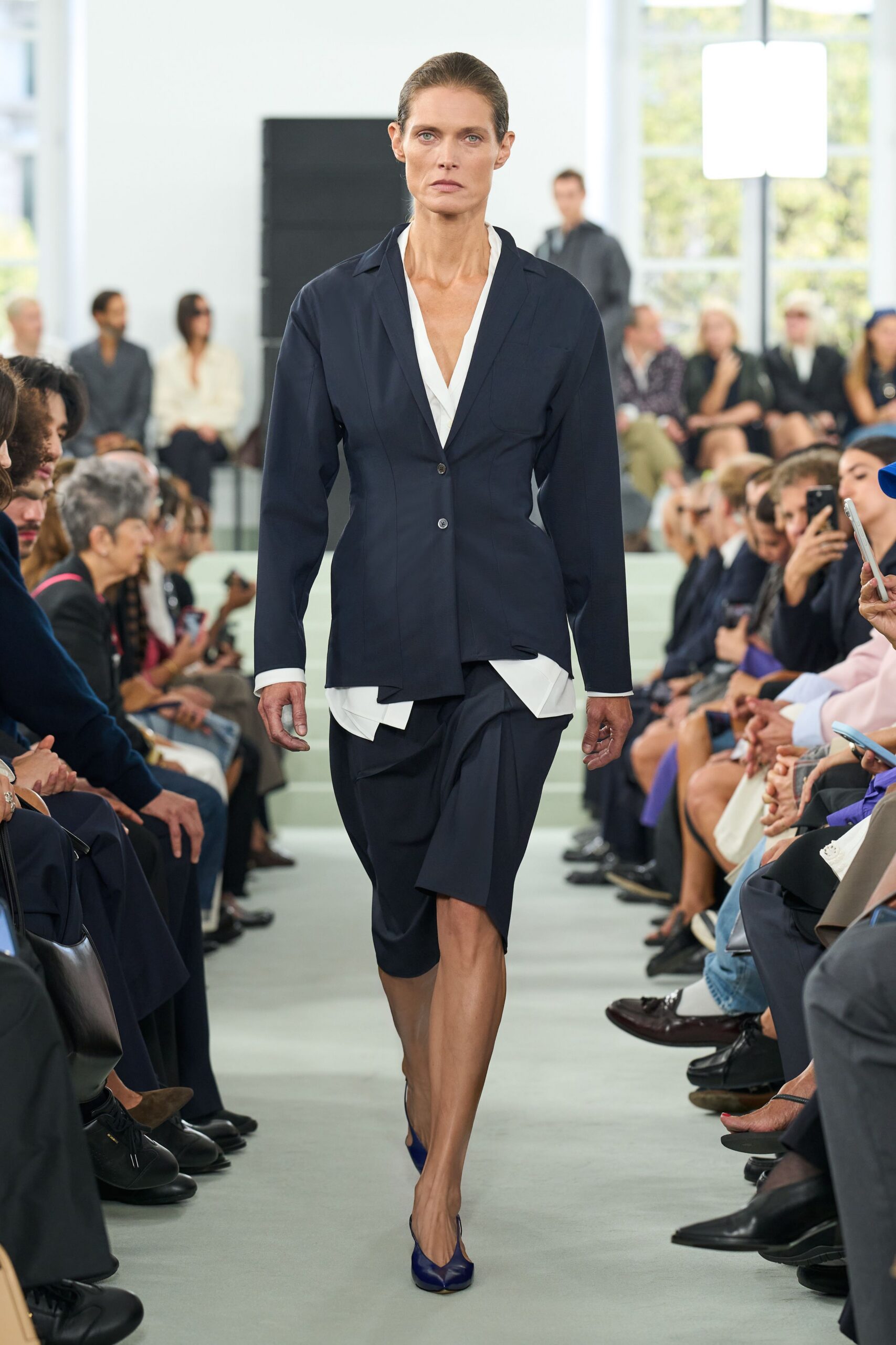 Ink navy blazer, one side jutting longer below the single-button front. White V-neck blouse, cuffs and hem peeking past the jacket. Matching wrap skirt draped into a soft knot over one thigh. Cobalt leather pumps with a sharply pinched vamp. Early meeting in a glass office before the coffee arrives.
