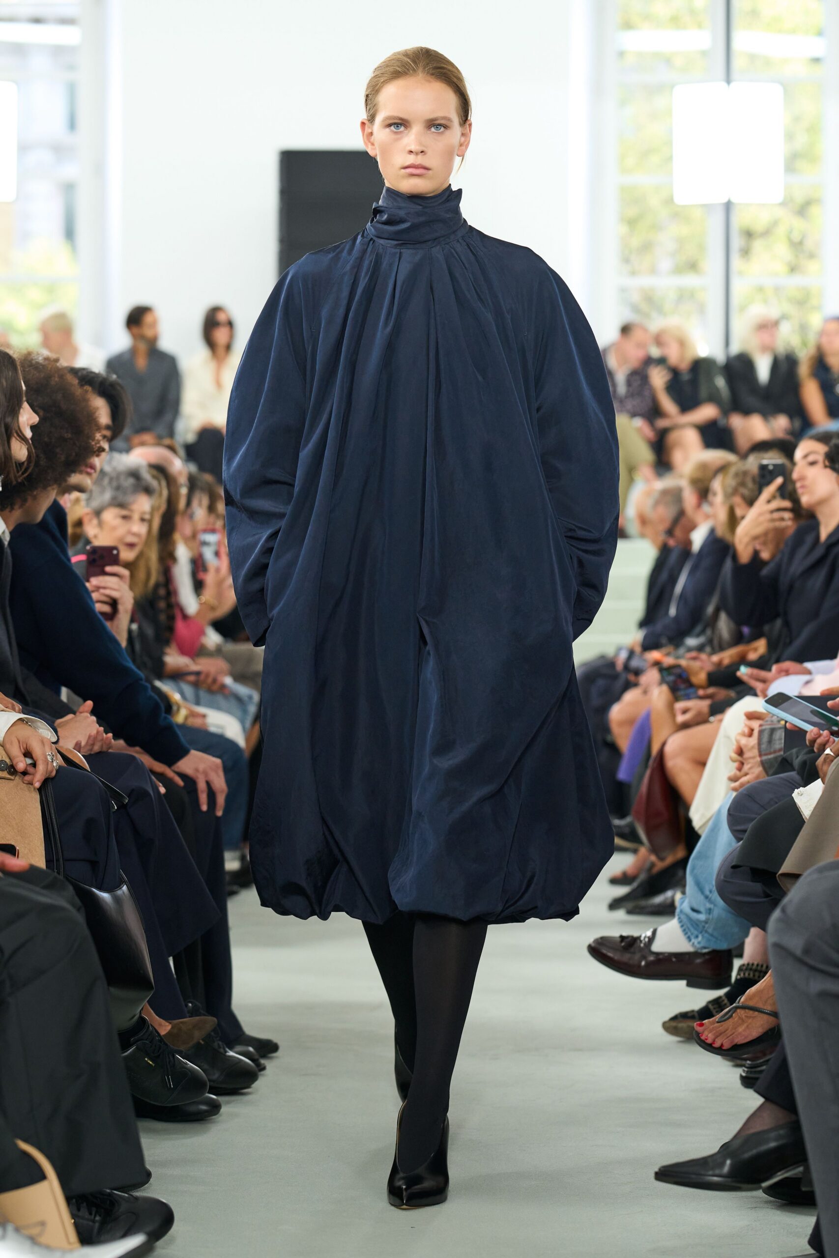 Ink-dark taffeta cocoon dress, gathered high at the throat and ballooned at the hem. Opaque black tights. Black split-toe pumps. Crossing a silent museum atrium before the doors open.