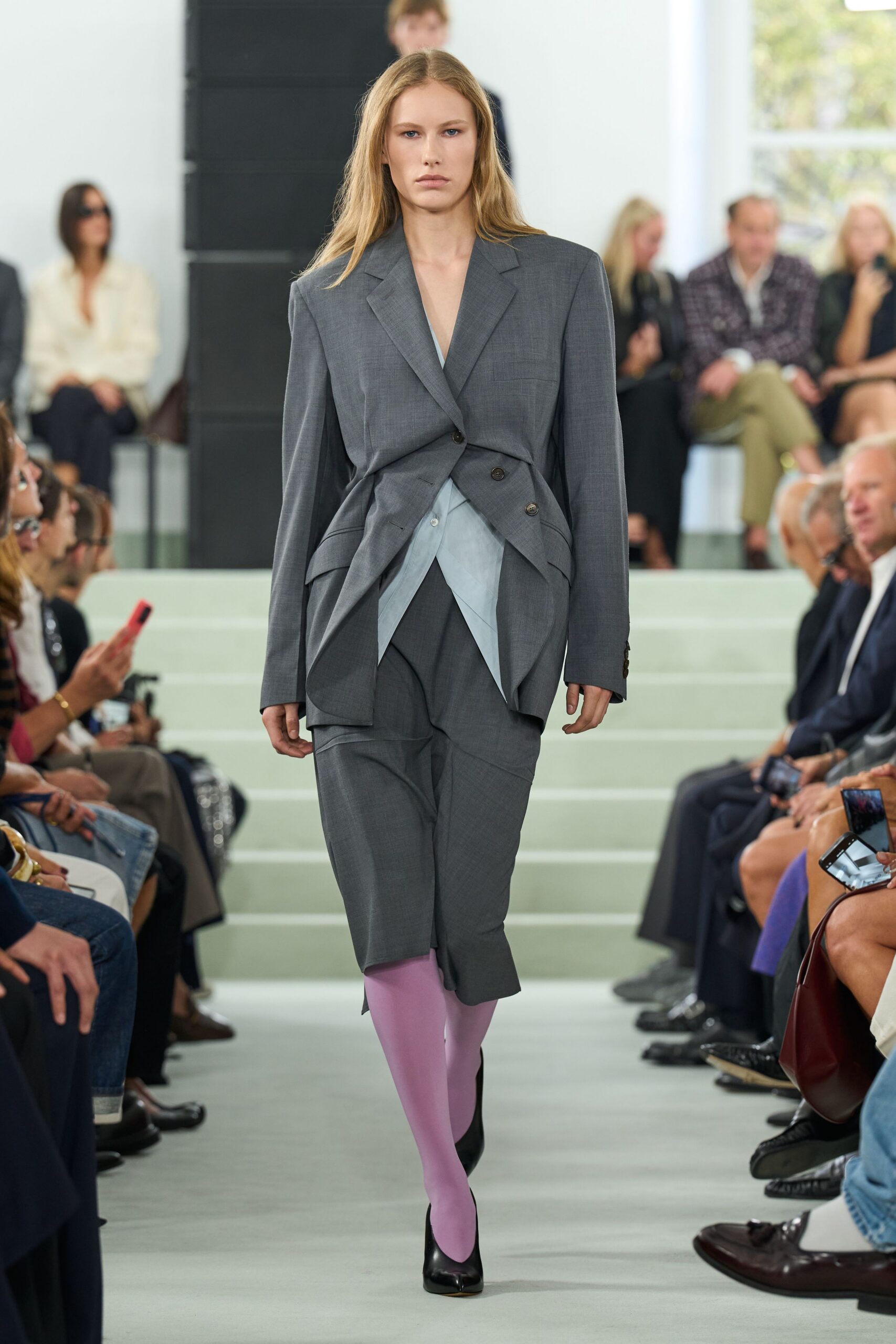 Grey suiting blazer twisted inward, one dark button catching the waist. Pale blue shirting points out beneath in two long triangular flaps. Matching skirt draped low and askew, slit opening over one shin. Lilac opaque tights. Black leather pumps, squared at the throat. Crossing a civic atrium before the hearing starts.