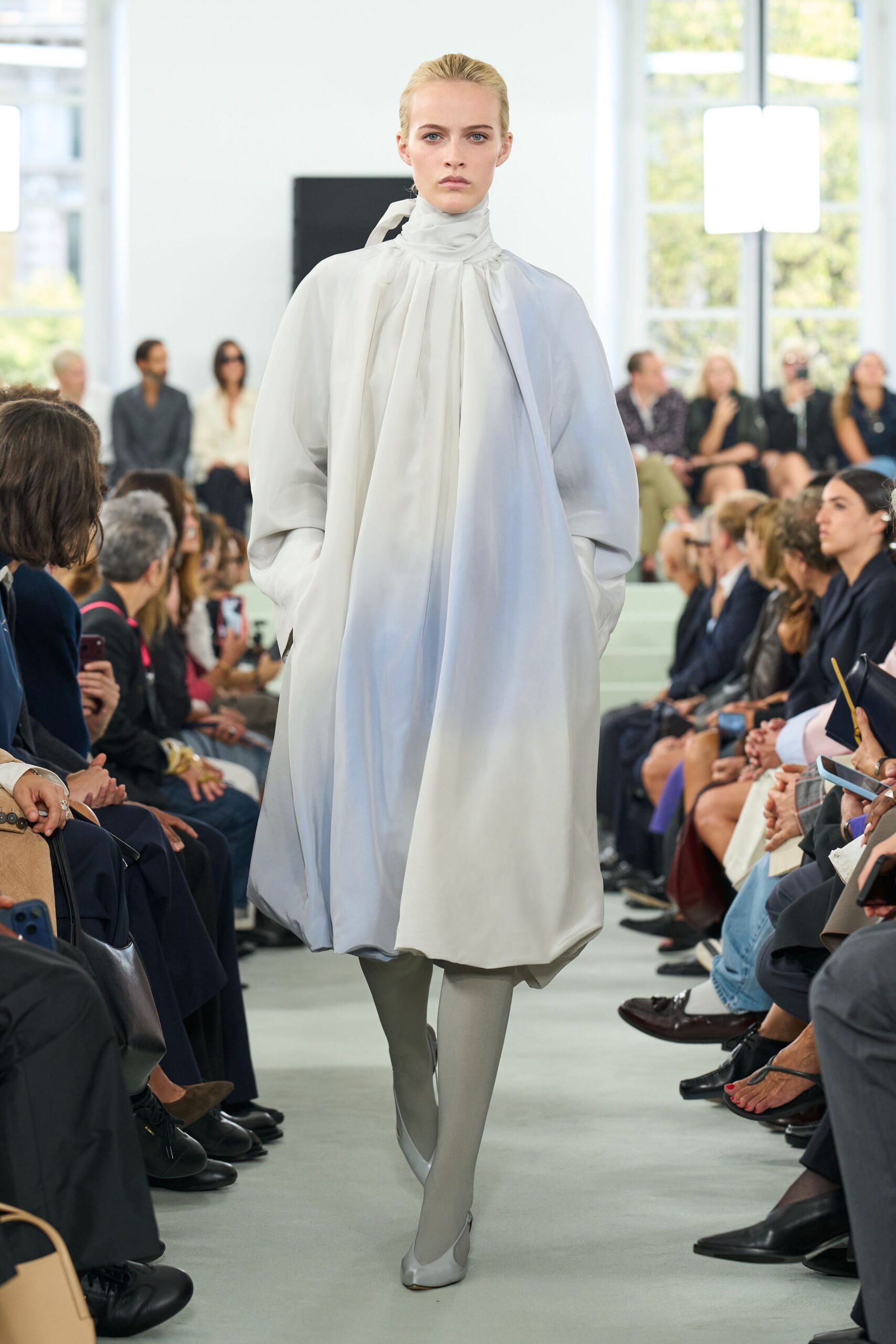 Pale dove silk dress with a wrapped neck and ballooned hem. Opaque pearl tights. Matching satin flats with a squared, tucked-looking toe. Crossing a museum atrium before the rain starts.