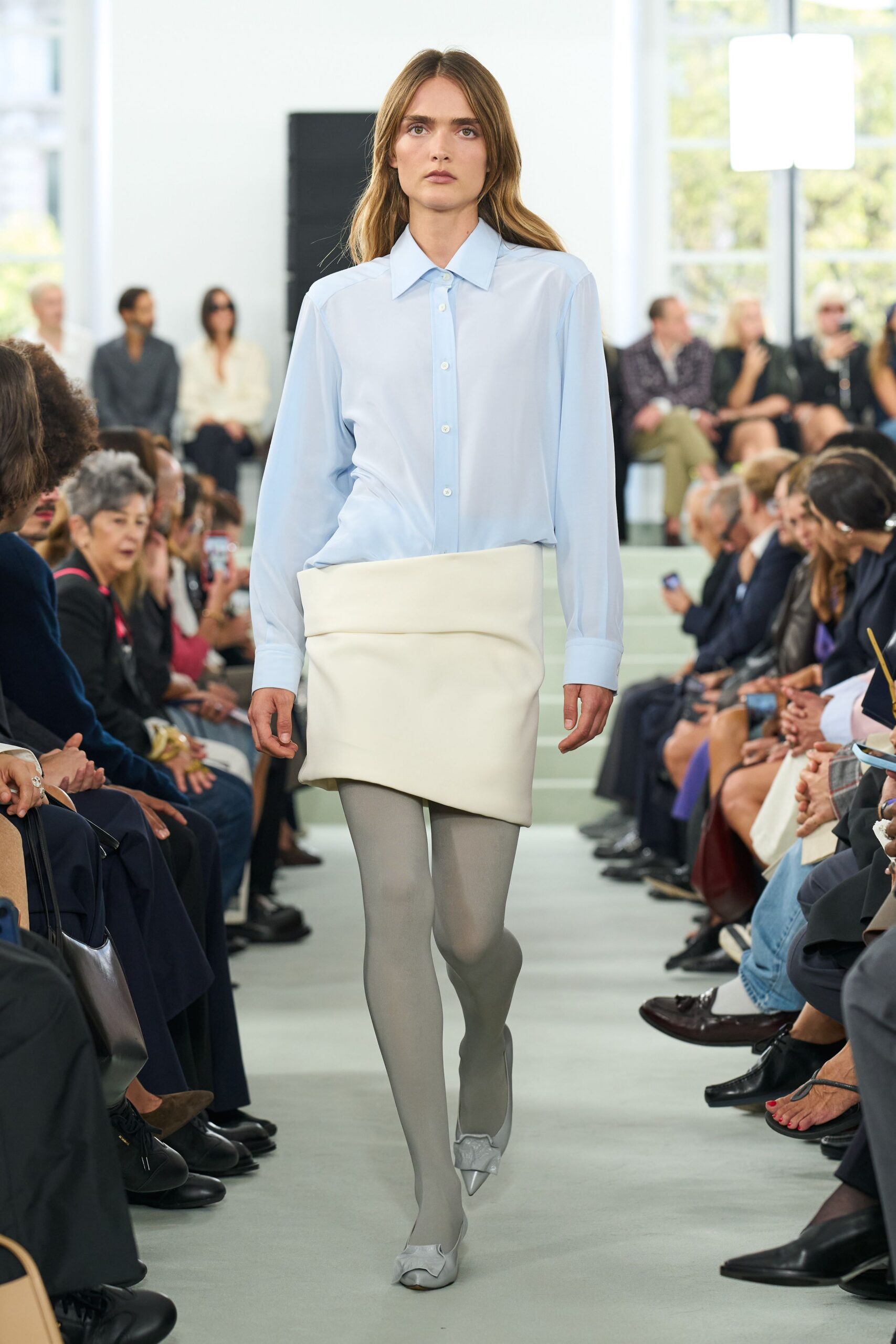 Pale blue shirt in fluid crepe, long cuffs and a pointed collar. Ivory mini skirt built like a folded cushion, rounded at the hem. Fine dove-grey tights. Silver pumps with soft gathered bows over the toe. Morning presentation in a glass house, before the coffee cart opens.