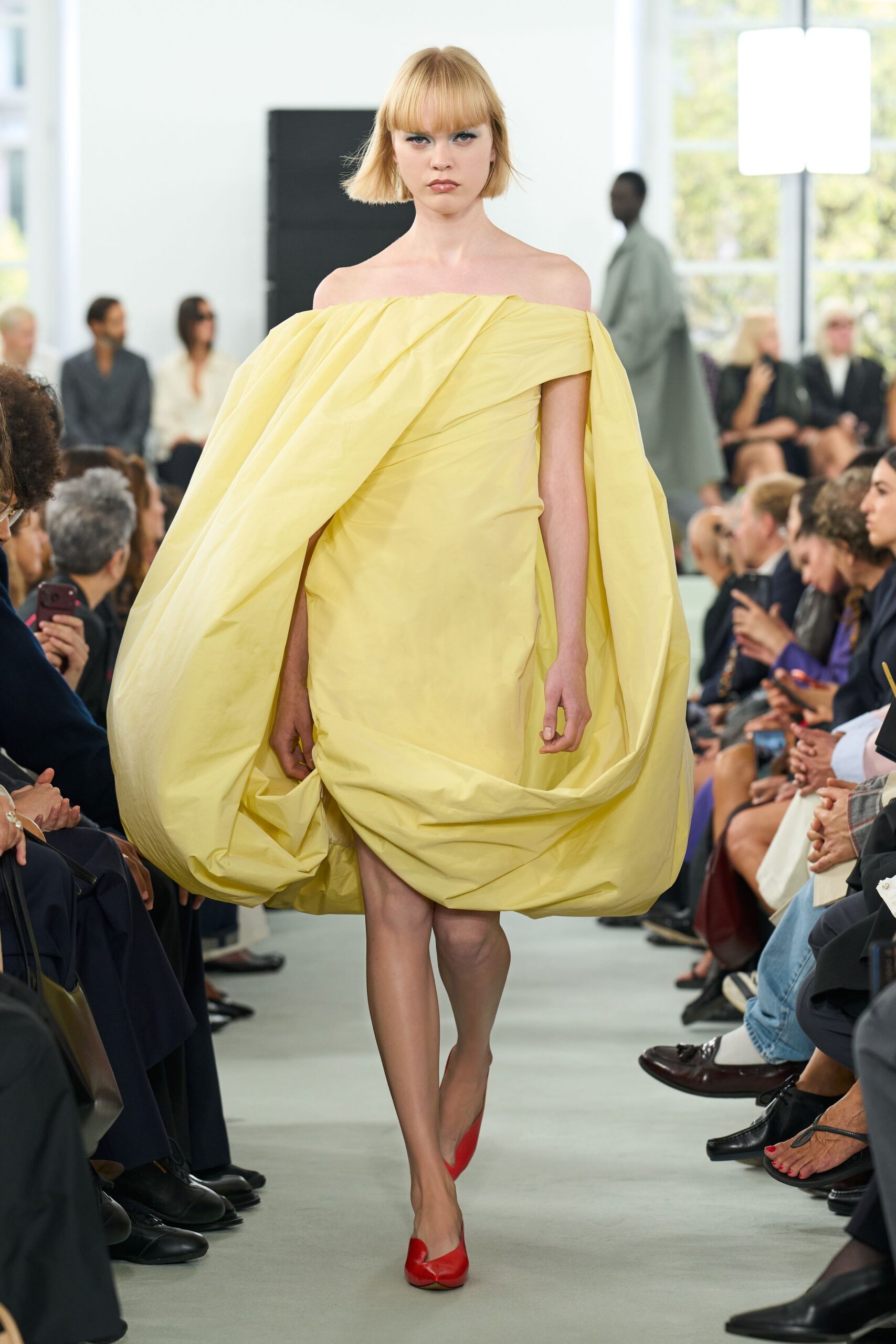 Butter-yellow taffeta dress with an off-shoulder neckline and enormous cocooned side drapes. Red leather pumps with a sharply split vamp. Crossing a marble foyer with florist paper still tucked under one arm.