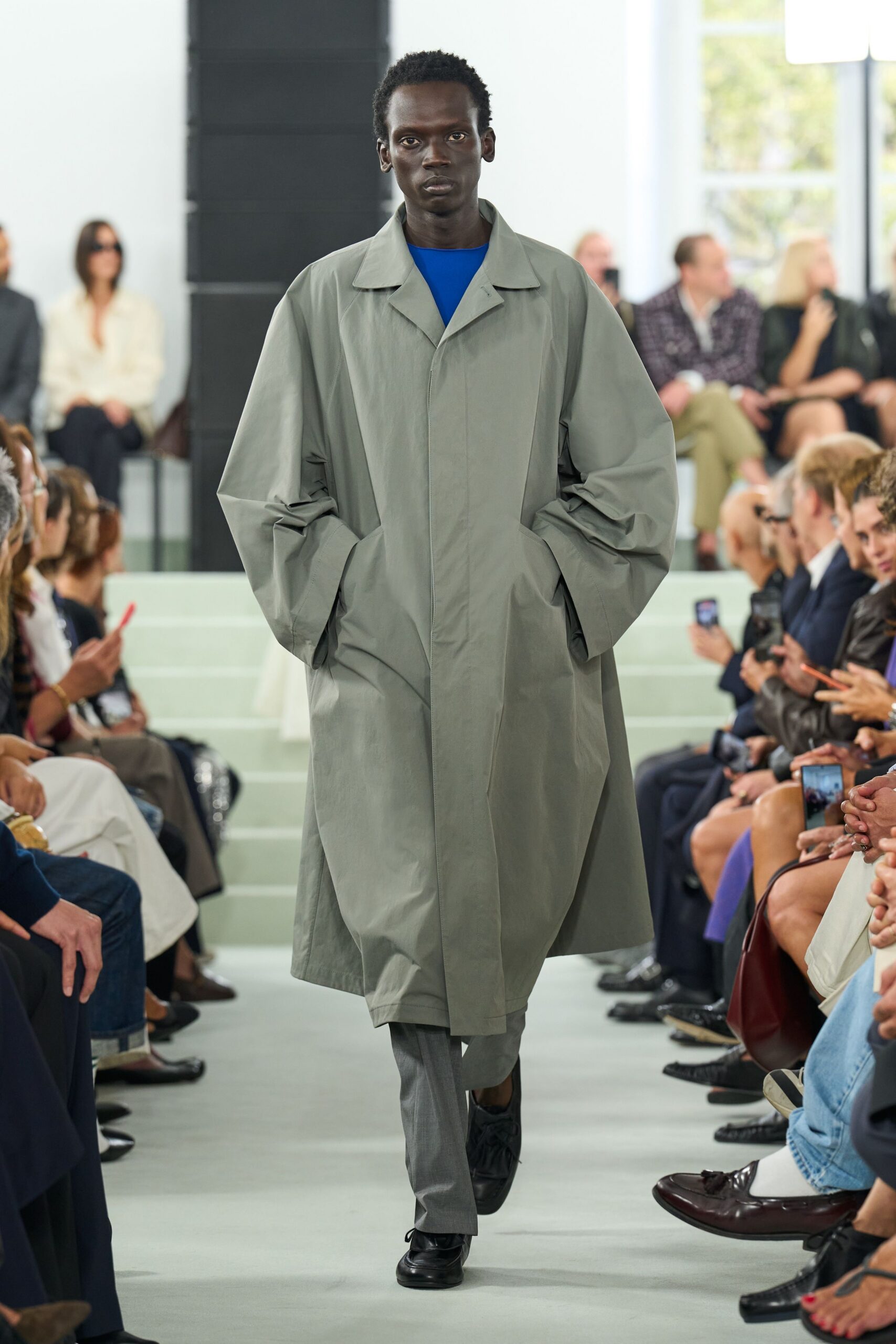 Pale sage mac in crisp cotton, sleeves ballooning above slashed pockets. Electric blue crewneck flashing cleanly at the collar. Grey tailored trousers with a narrow straight break. Black leather derbies with gathered apron fronts. Crossing a windy plaza before the office towers fill up.
