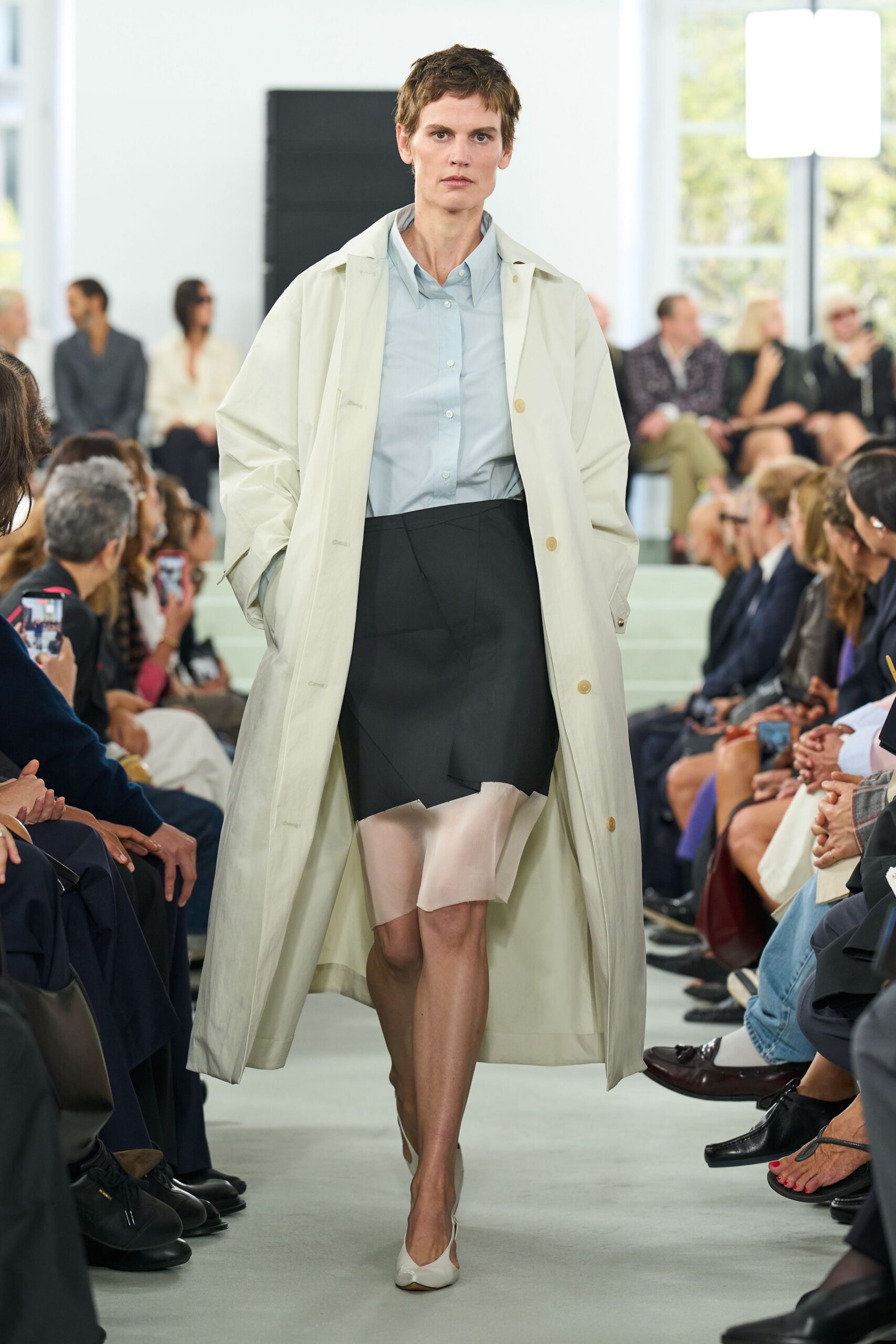 Long chalk trench coat with pale horn buttons and dropped shoulders. Powder-blue cotton shirt, collar slightly crumpled under the neckline. Black wrap-front skirt over a blush satin underlayer cut unevenly below. Milk-white pumps with a low vamp. Walking into a municipal atrium early, paperwork tucked under one arm.