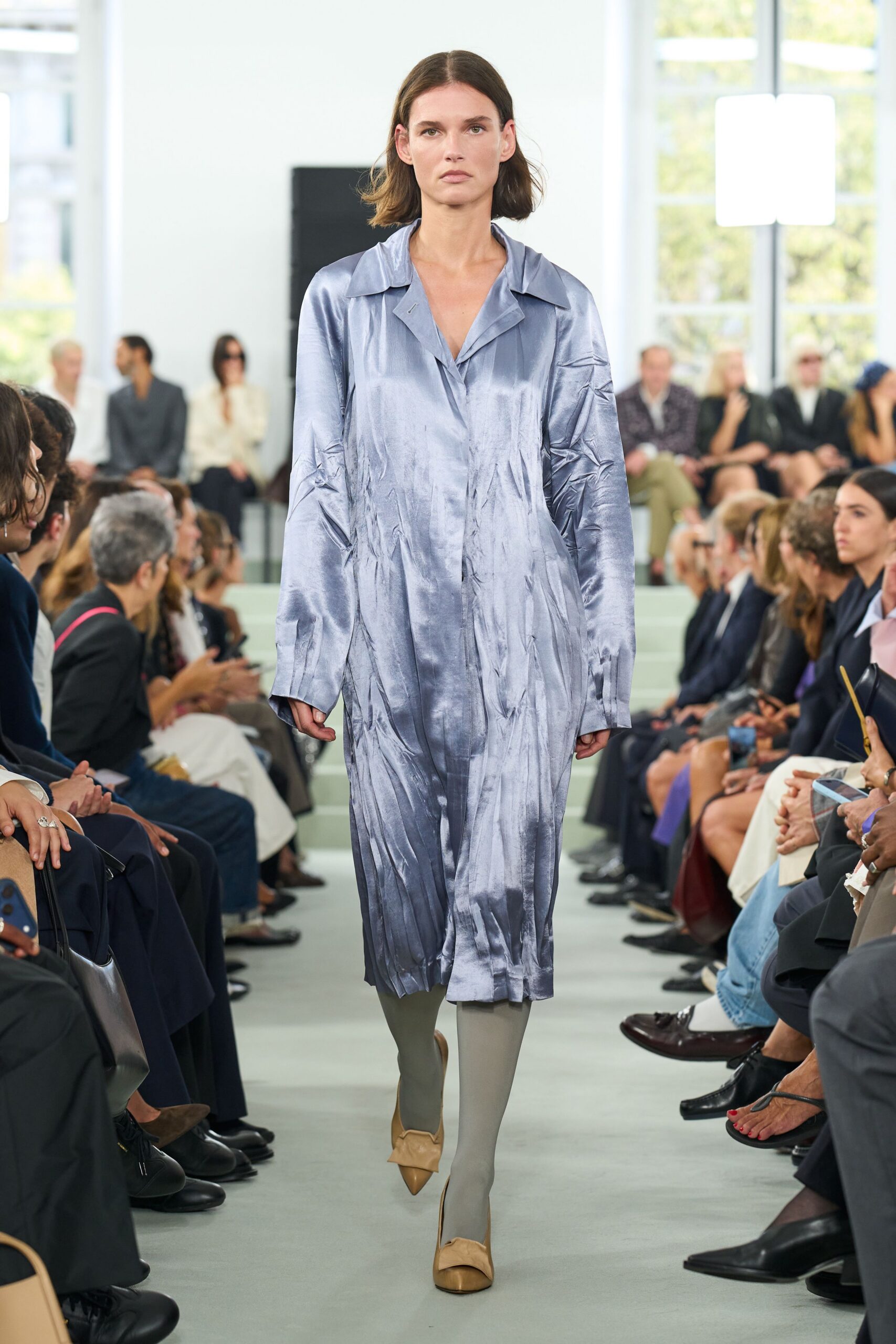 Steel-lilac satin shirt dress, crushed into long vertical creases and soft glare. Pale sage opaque tights. Tan leather pumps with a folded bow pinched over the toe. Morning presentation in a glass office where the air-conditioning never lets up.
