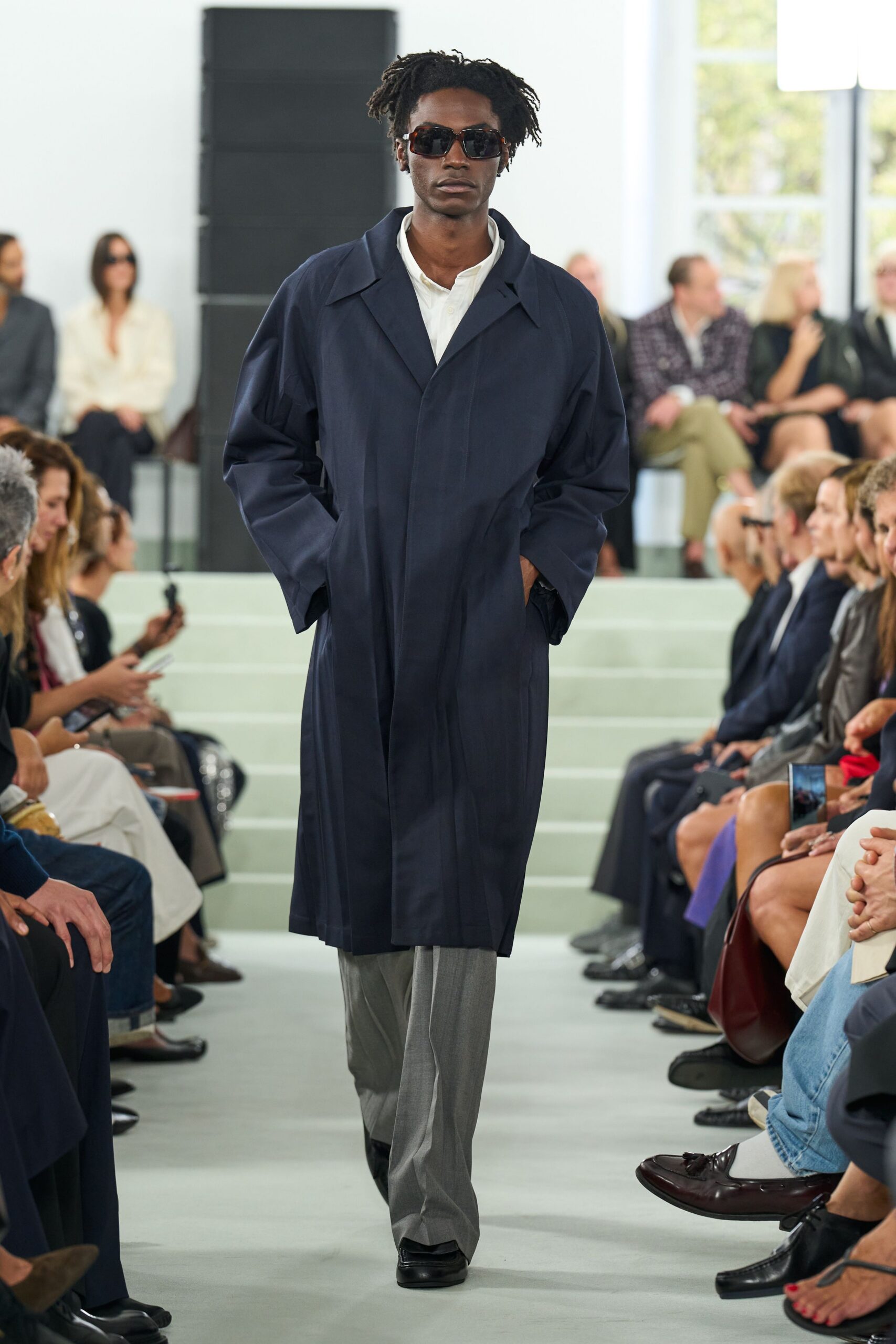 Dark tortoiseshell sunglasses with narrow rectangular lenses. Ink navy overcoat in crisp cloth, cut long with dropped raglan sleeves. White band-collar shirt, top button left open. Pale grey trousers with a sharp front crease. Black square-toe shoes. Crossing a quiet corporate plaza before the revolving doors unlock.