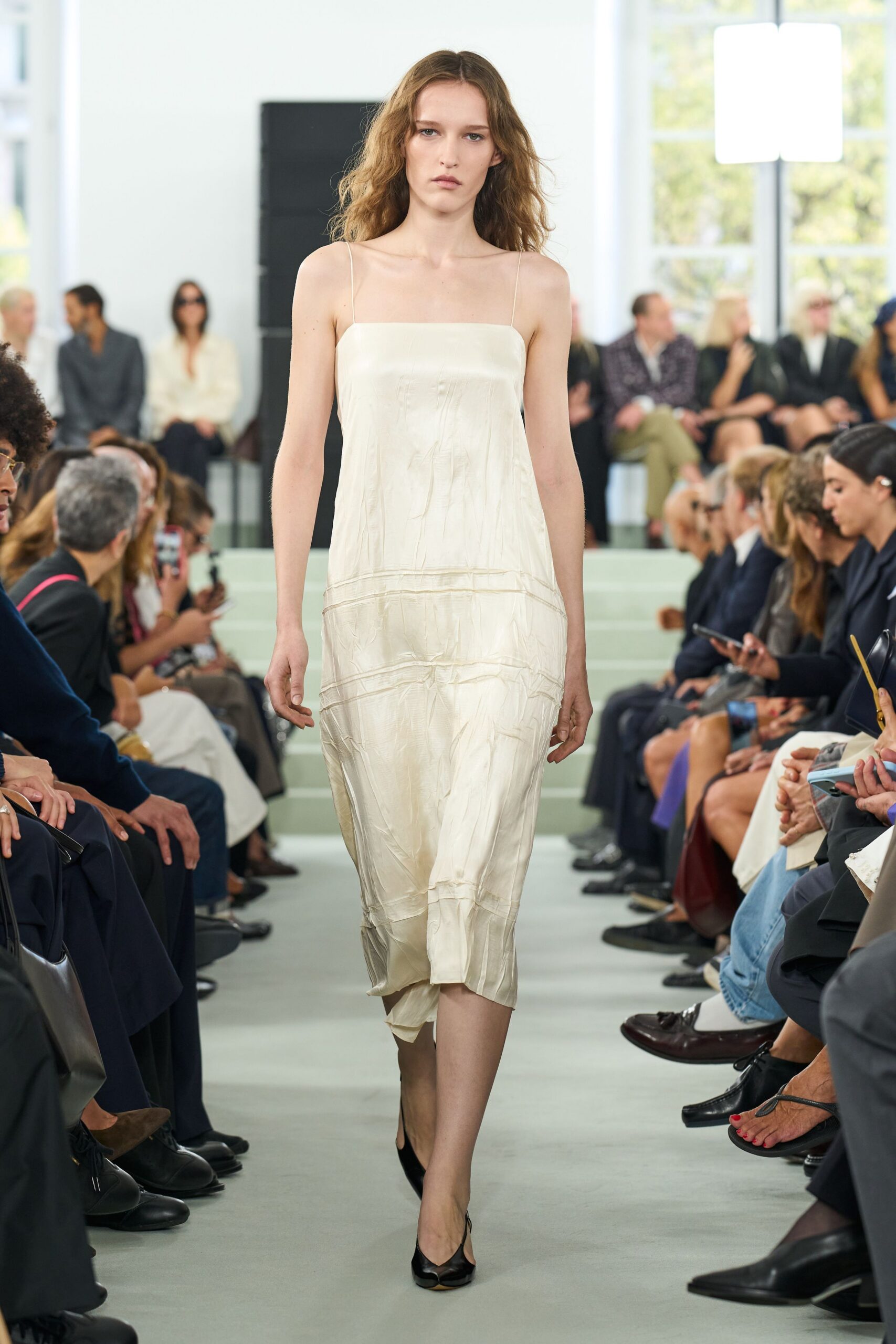 Ivory satin slip dress, wrinkled through the body with horizontal seam bands. Black split-toe slingback heels. Morning after the wedding, carrying your shoes down a cold hotel corridor.