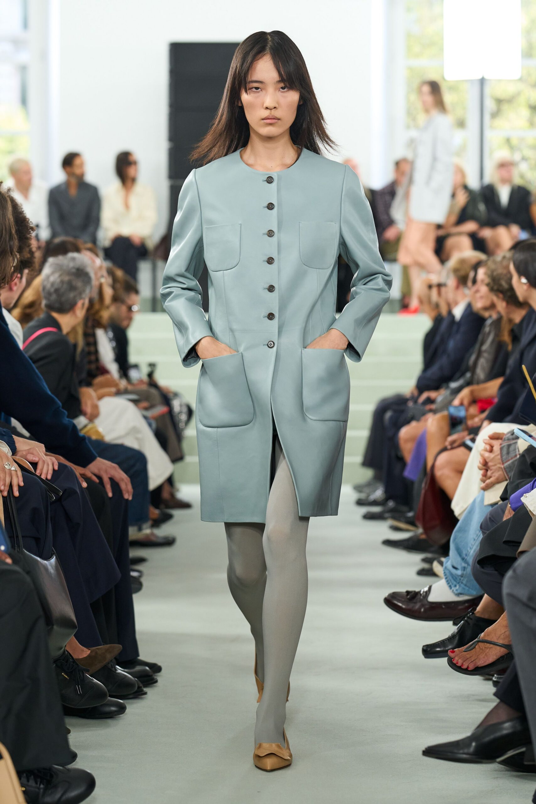 Powder-blue leather coatdress with rounded patch pockets and dark horn buttons. Pale grey opaque tights. Camel pumps with a softly squared toe. Walking into a municipal atrium early, before the building warms up.