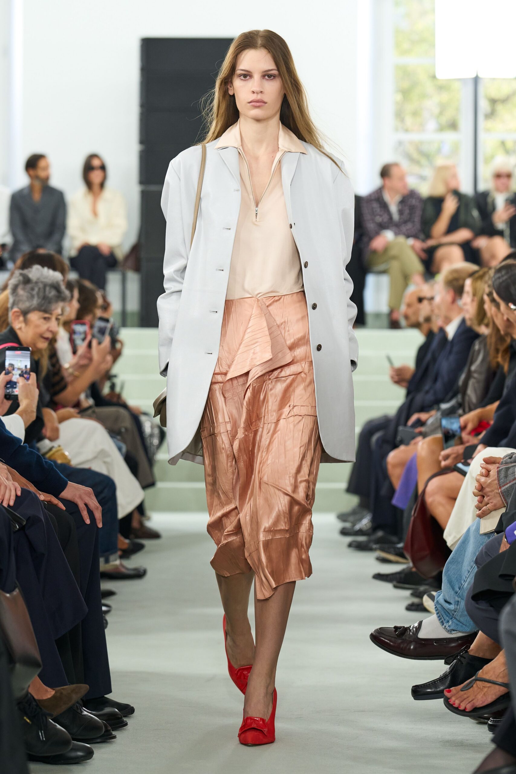 Pale grey coat with dark horn buttons and rounded cutaway hem. Blush satin zip-neck top, collar spread open at the throat. Coppery crinkled skirt wrapped in uneven cascading panels. Sheer taupe tights. Scarlet satin pumps with soft folded bows. Walking into a hotel atrium while the florist is still hosing the stone.