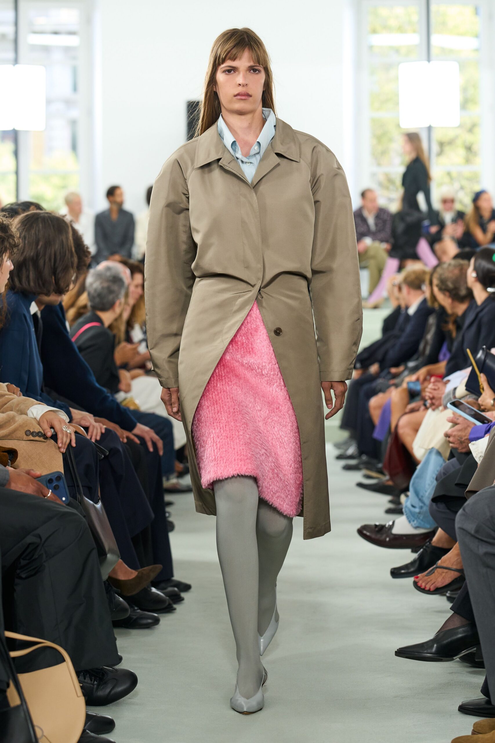 Taupe coat twisted shut at the waist, one brown button left exposed. Pale blue button-down peeking from the open collar. Shaggy pink skirt cut on a diagonal below the knee. Opaque dove-grey tights. Pointed grey pumps with low-cut vamps. Walking into the office on the first cold morning after a beach holiday.