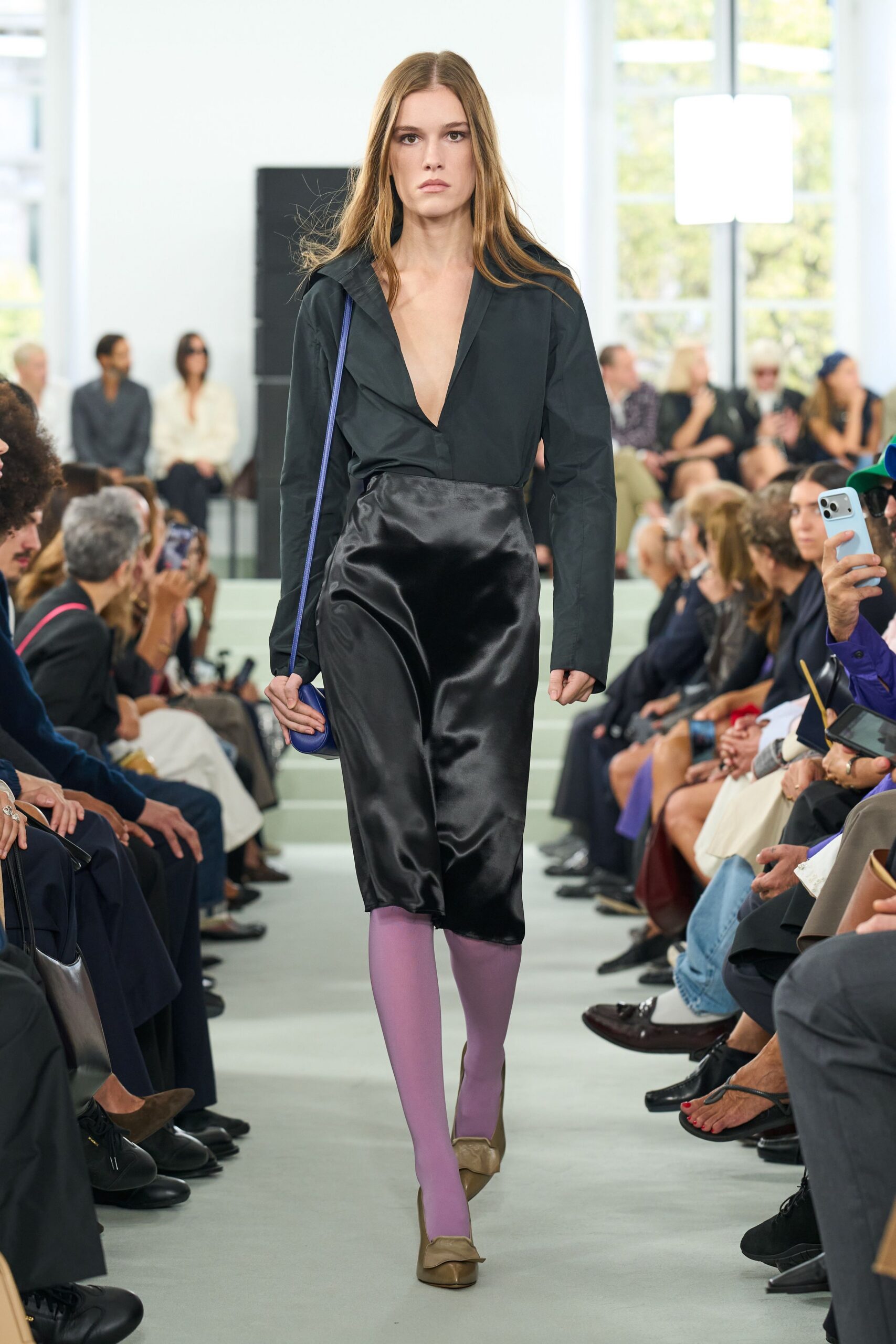 Black cotton blouse cut to a plunging V, collar spread open. Black satin midi skirt, pooled shine and a slight twist at hem. Cobalt leather pouch on a narrow strap. Lilac opaque tights. Taupe pumps with a softly folded apron toe. Crossing the hotel atrium early, before the conference badges come out.