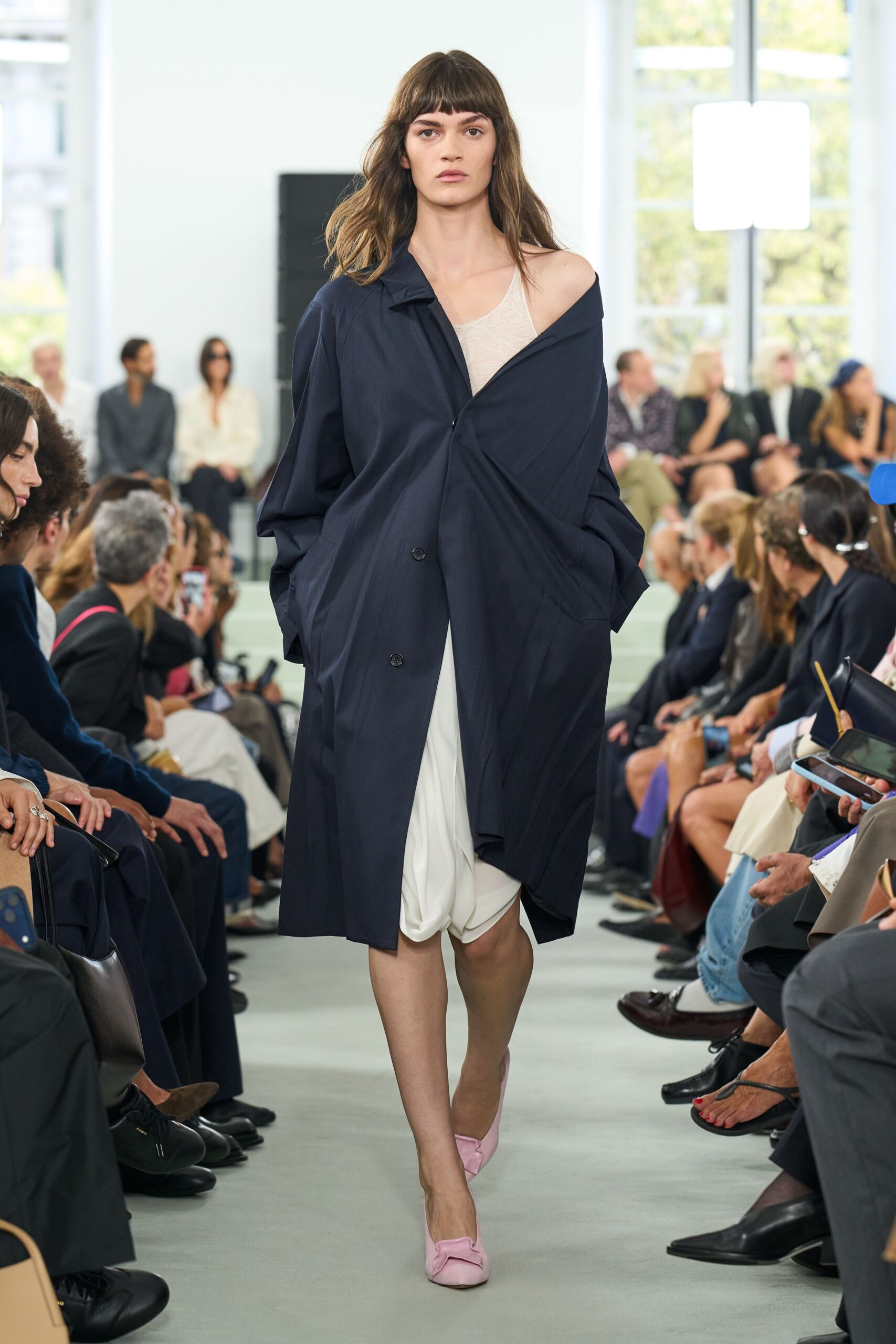 Oversized navy coat twisted off one shoulder, two dark buttons anchoring the drape. Thin ivory knit tank, sheer across the chest. White jersey skirt gathered into a soft pouch at the hem. Powder-pink flats with folded bow fronts. Whatever you pull on for a ferry crossing before the sun burns off.